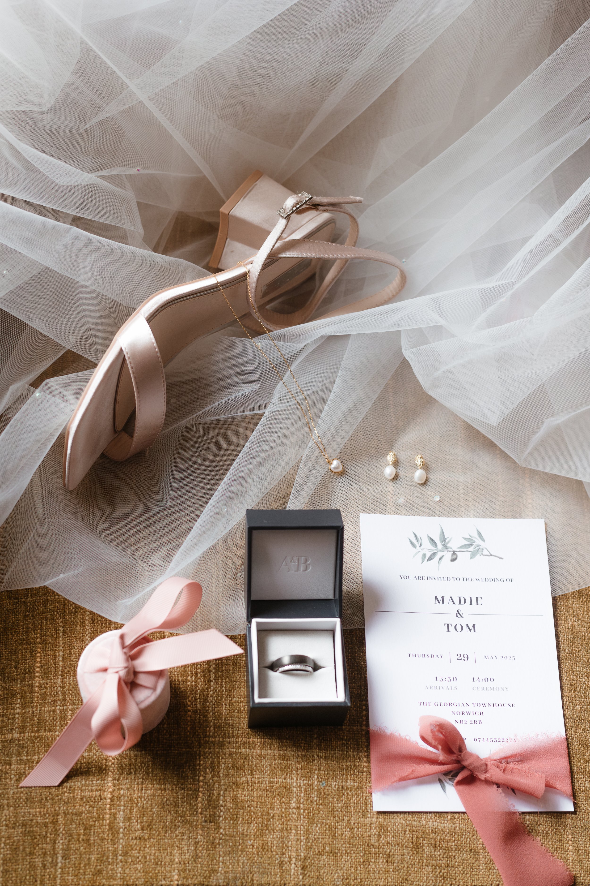 Wedding accessories including a pair of pearl drop earrings, a pearl necklace, a wedding invitation, wedding rings in a box, a pink wrapped gift, a pair of nude high-heeled shoes, and sheer white fabric on a brown surface.