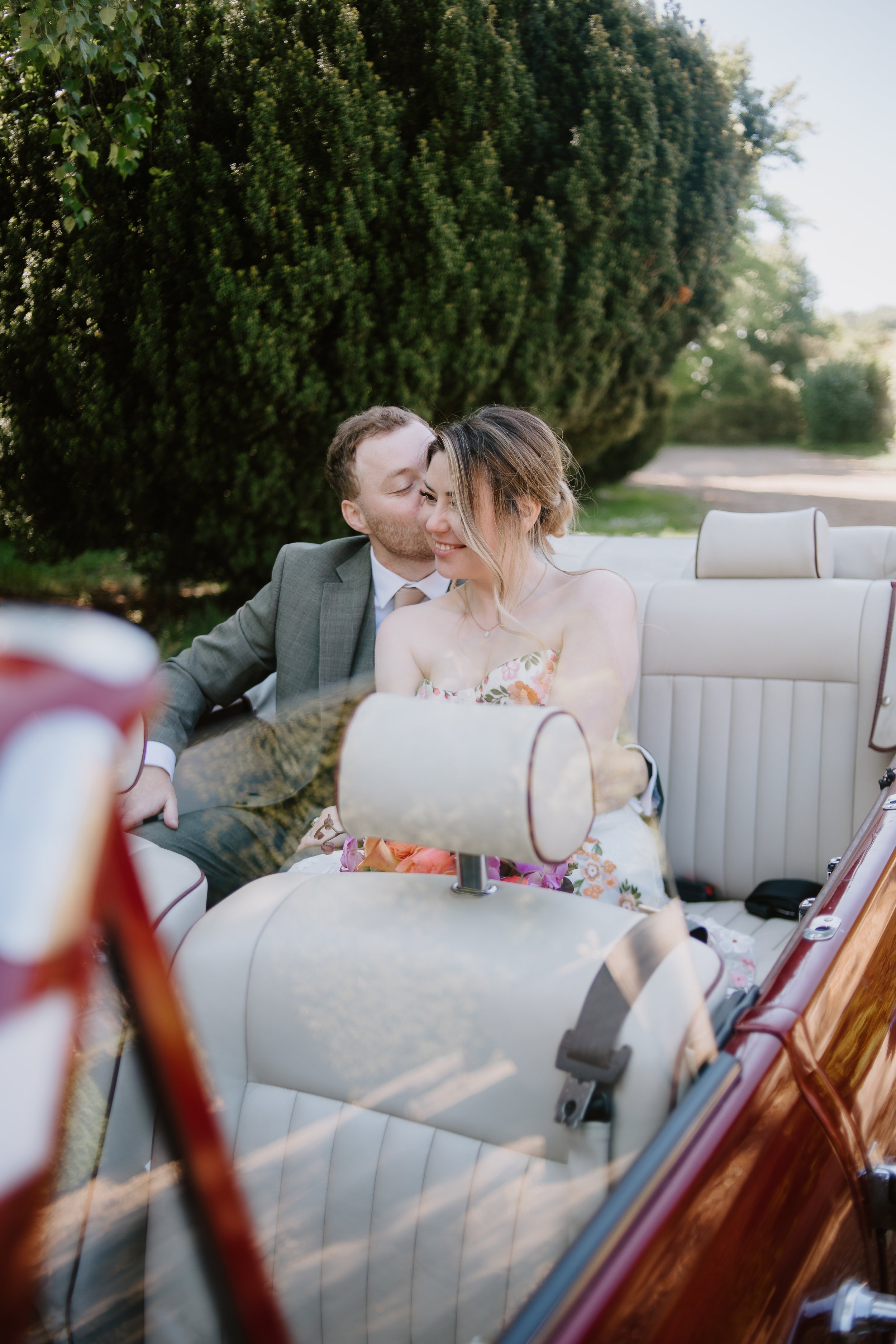 A couple dressed in wedding attire sharing a tender moment and smiling while sitting in a vintage convertible car outside surrounded by greenery.