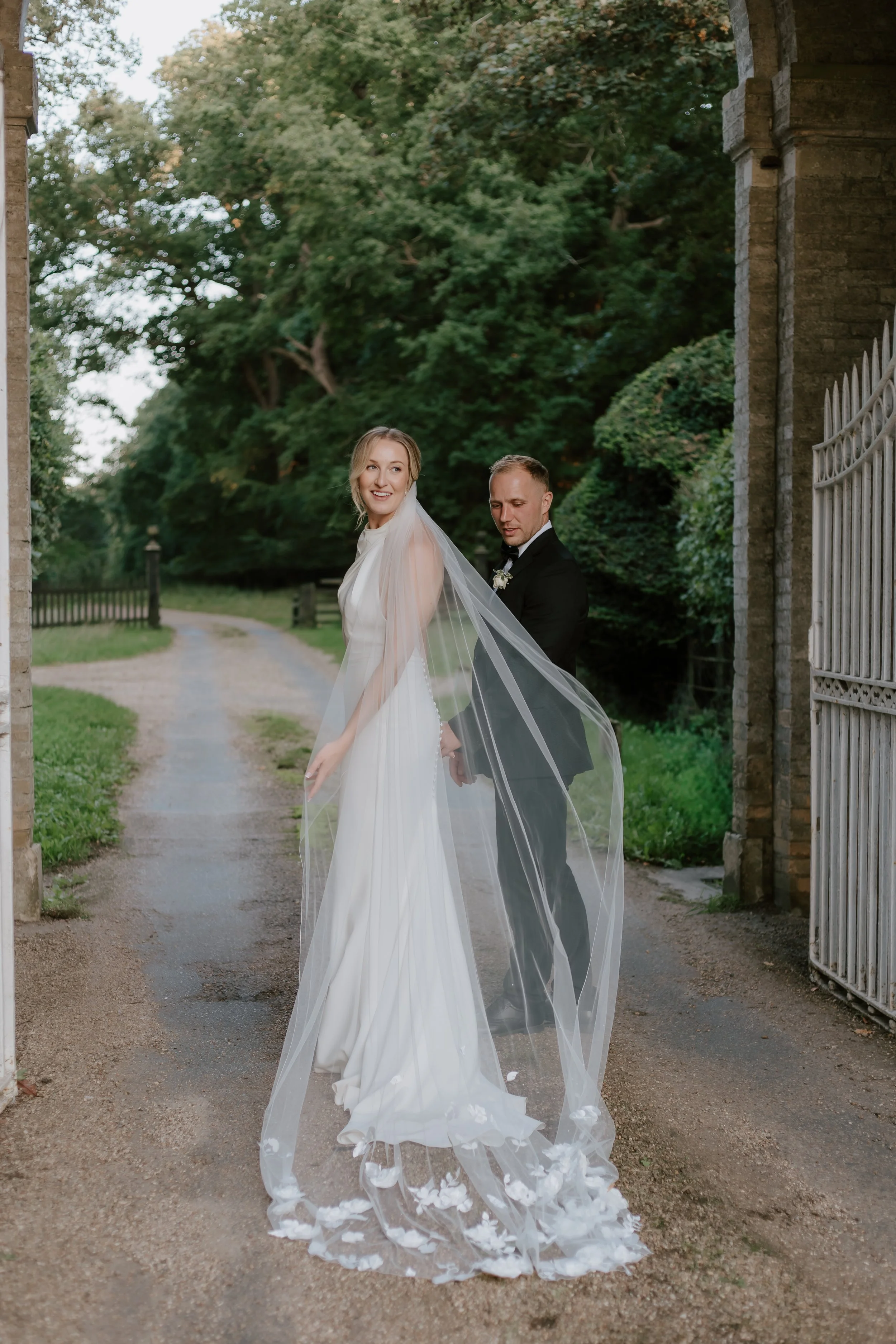 Romantic and natural wedding photographer Suffolk and Norfolk – bride and groom holding hands and smiling outdoors by stone archway and green trees.