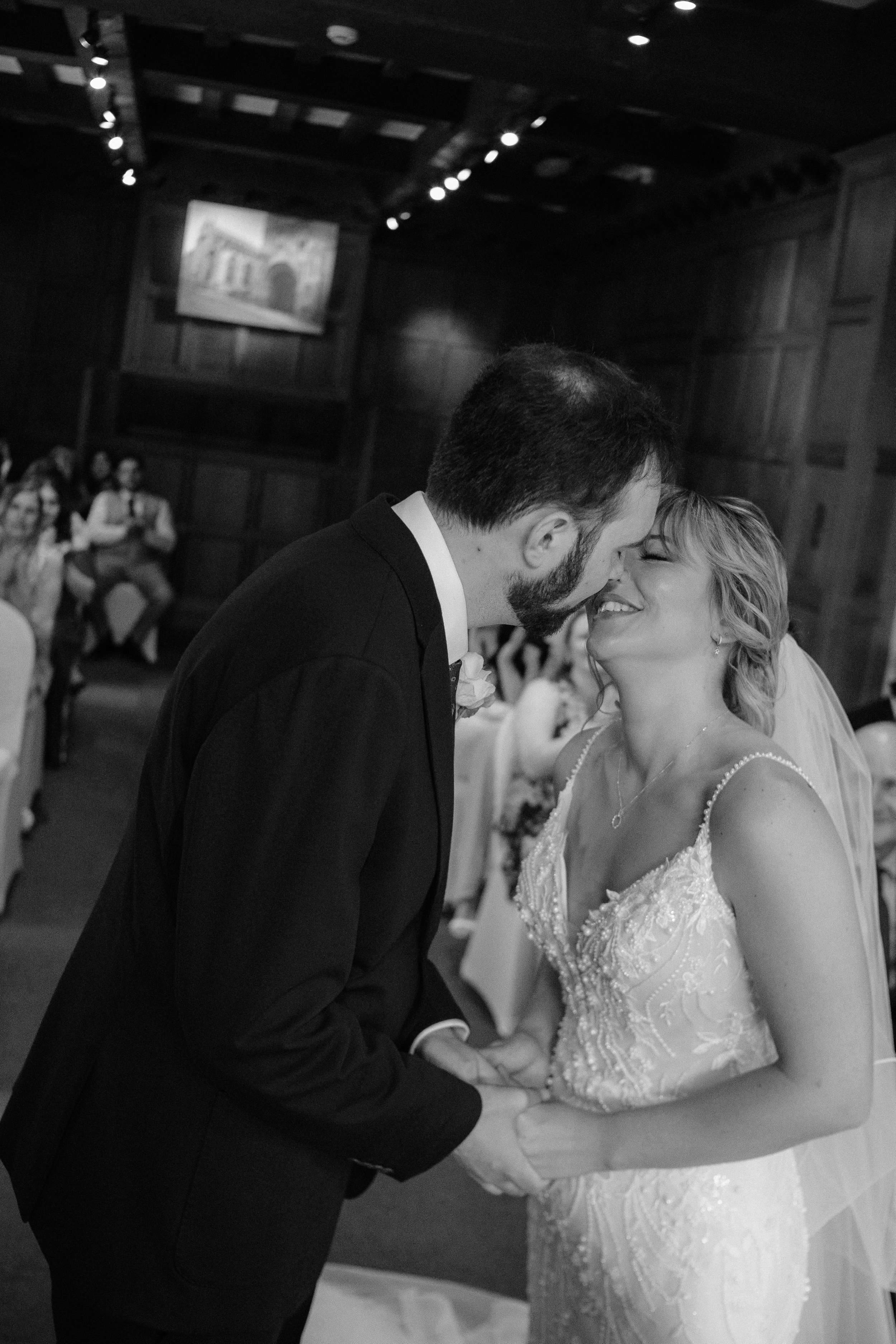 A black and white photograph of a wedding ceremony showing a bride and groom sharing a kiss, holding hands, in a wood-paneled room with seated guests in the background.