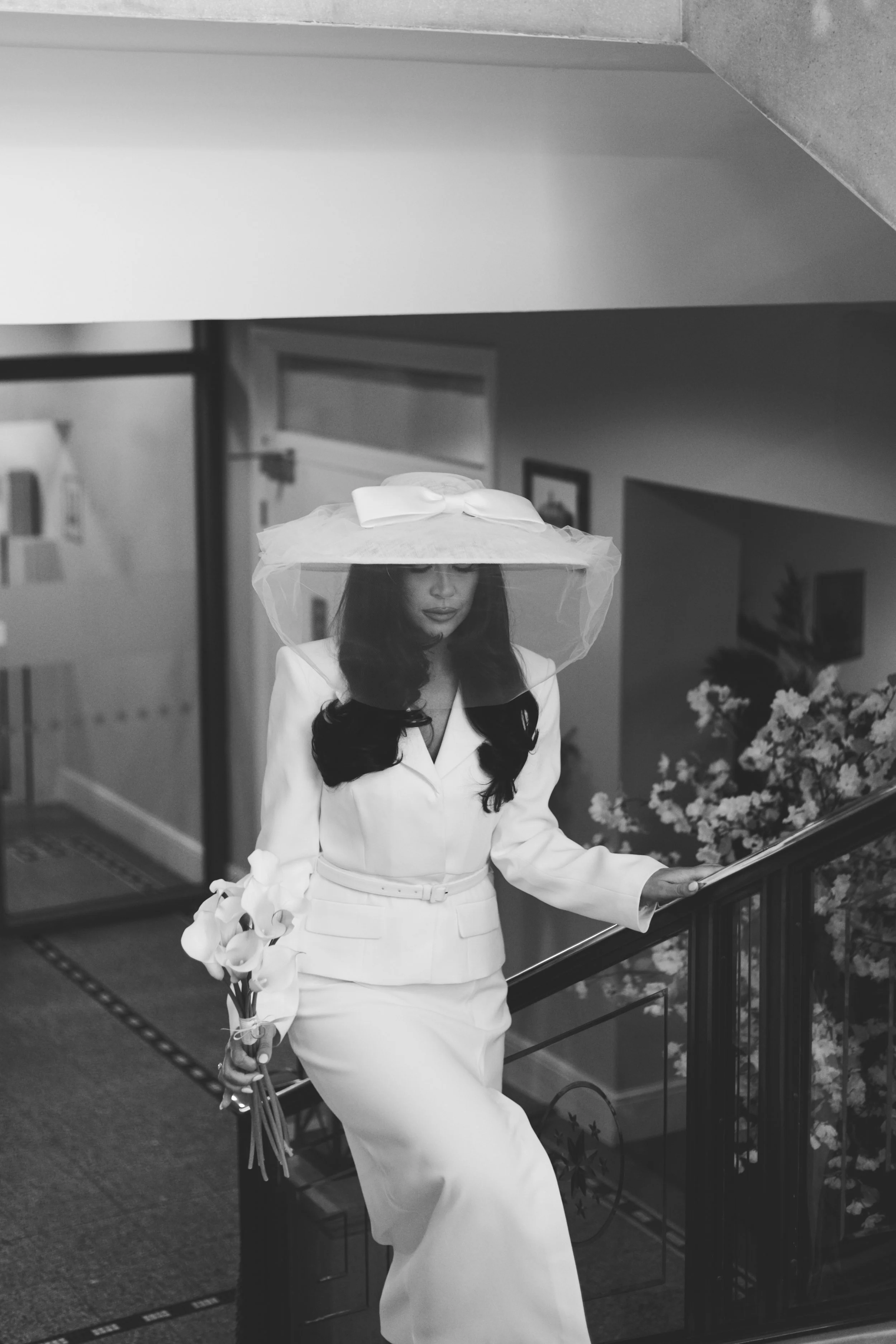 A woman dressed in an elegant white outfit with a large hat and veil, holding a bouquet of calla lilies, walking down a staircase.