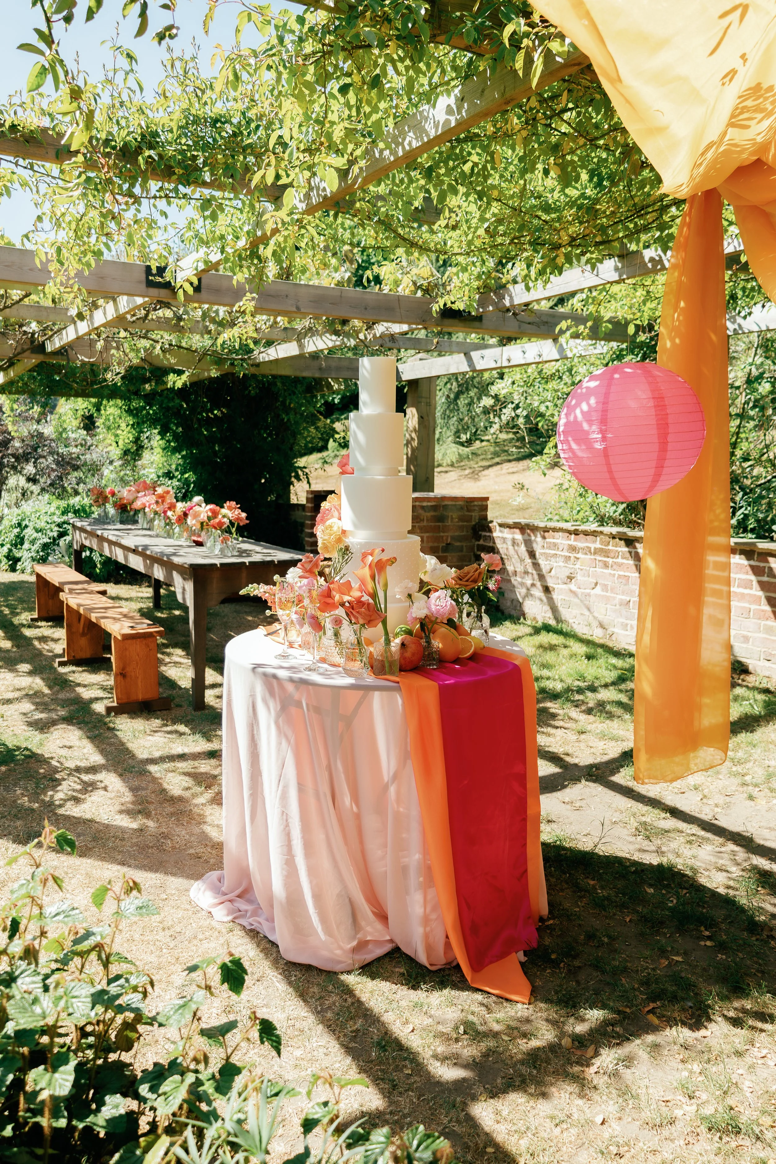 Decorated outdoor celebration table with a tiered cake, flowers, and fruit under a pergola with sunlight and colorful paper lanterns.