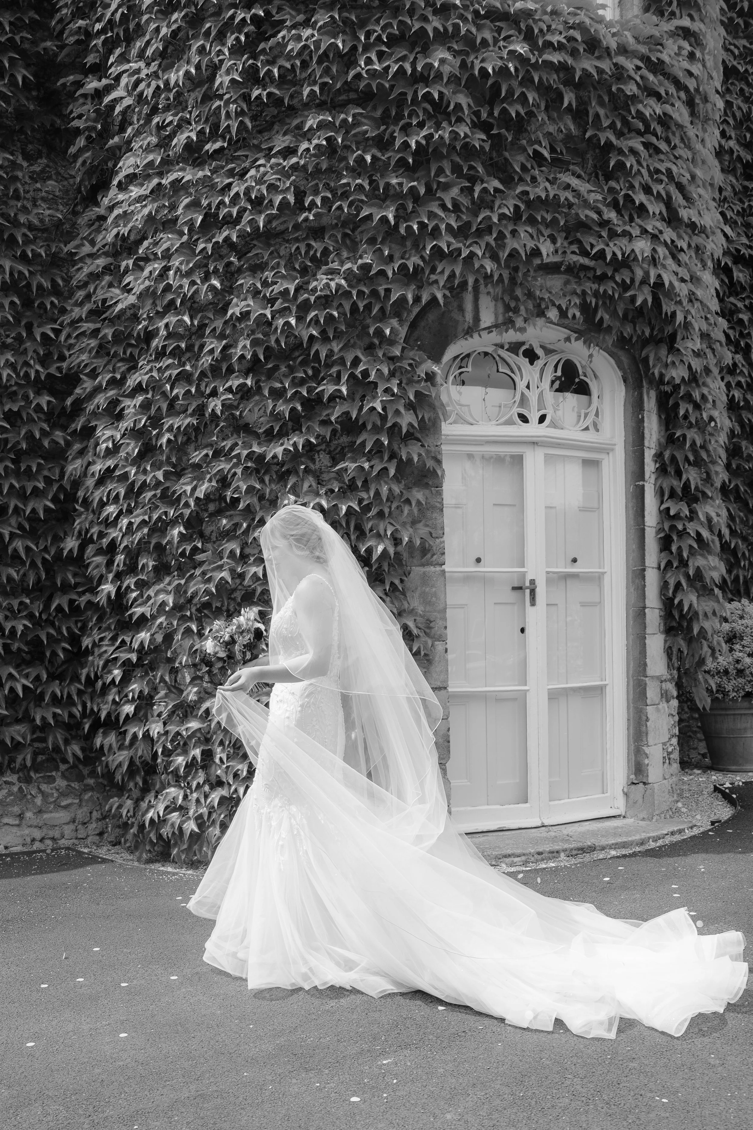 Black and white photo of a bride in a wedding gown and veil, standing outdoors in front of a ivy-covered wall and a white double door.