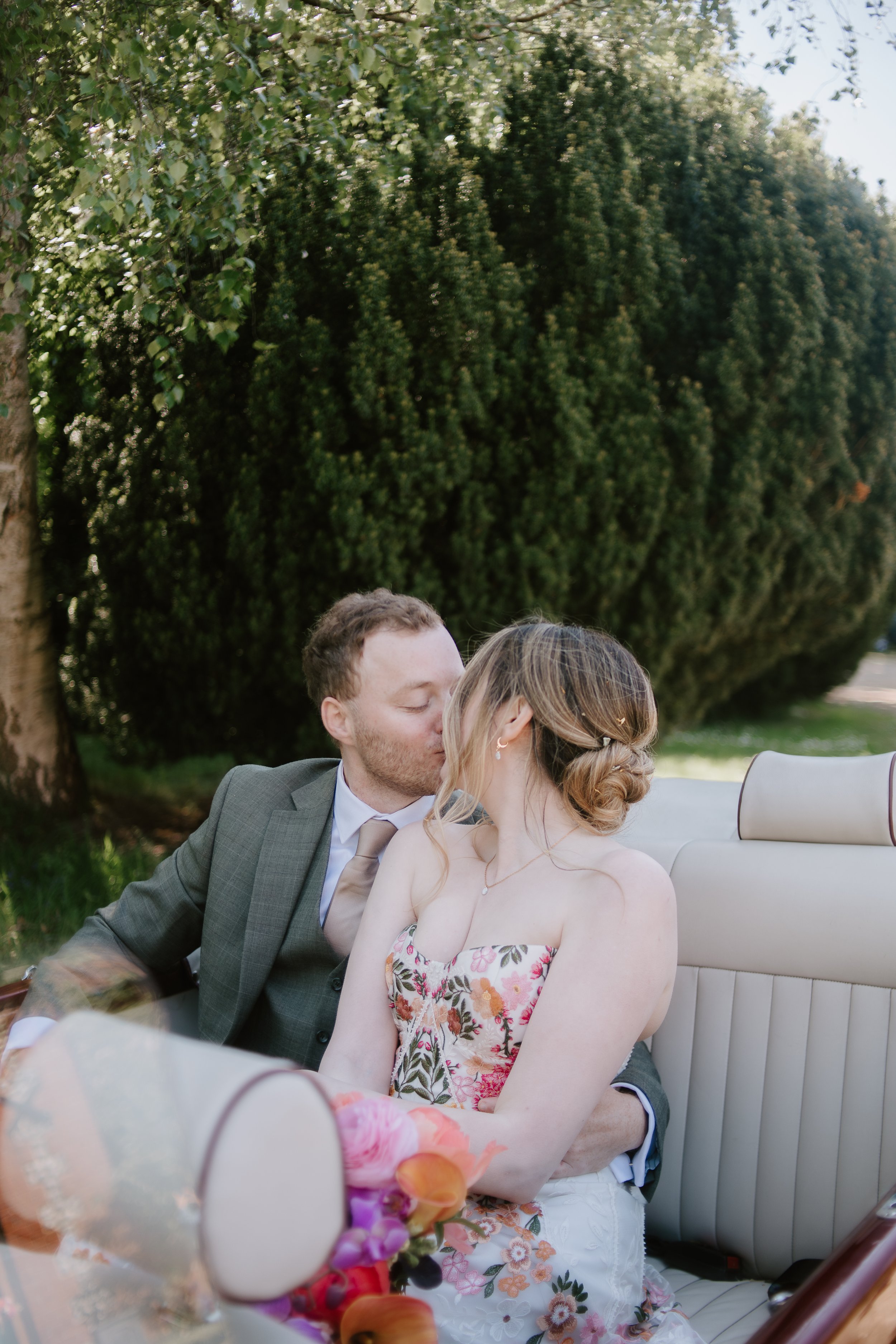 A couple is sitting in a vintage car, sharing a kiss outdoors during daytime. The man is wearing a gray suit, and the woman is in a strapless floral dress with a bouquet of flowers in the foreground. Green trees are visible in the background.
