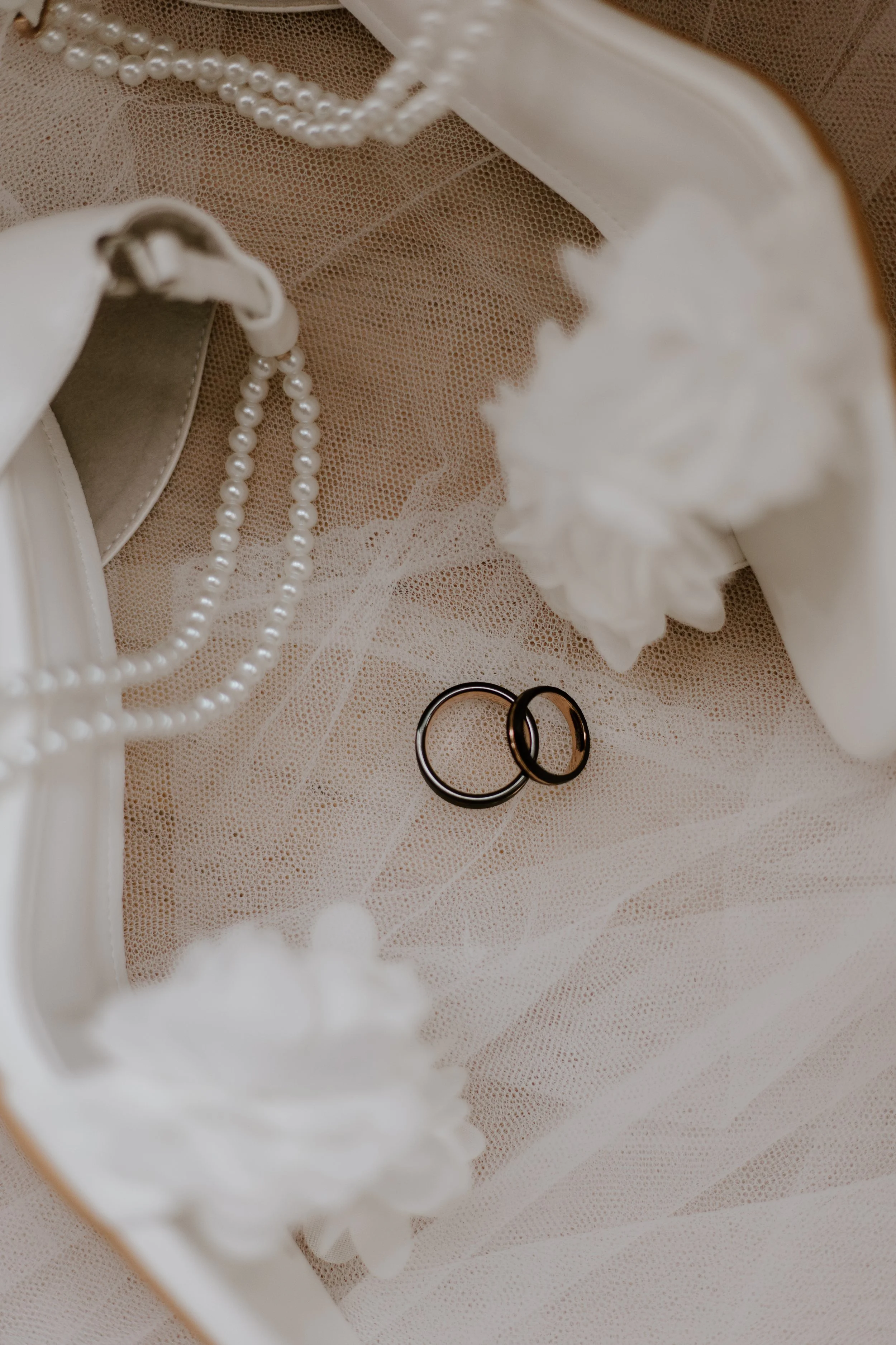 Two wedding rings placed on a decorative white veil or fabric surrounded by lace, pearls, and white flowers.
