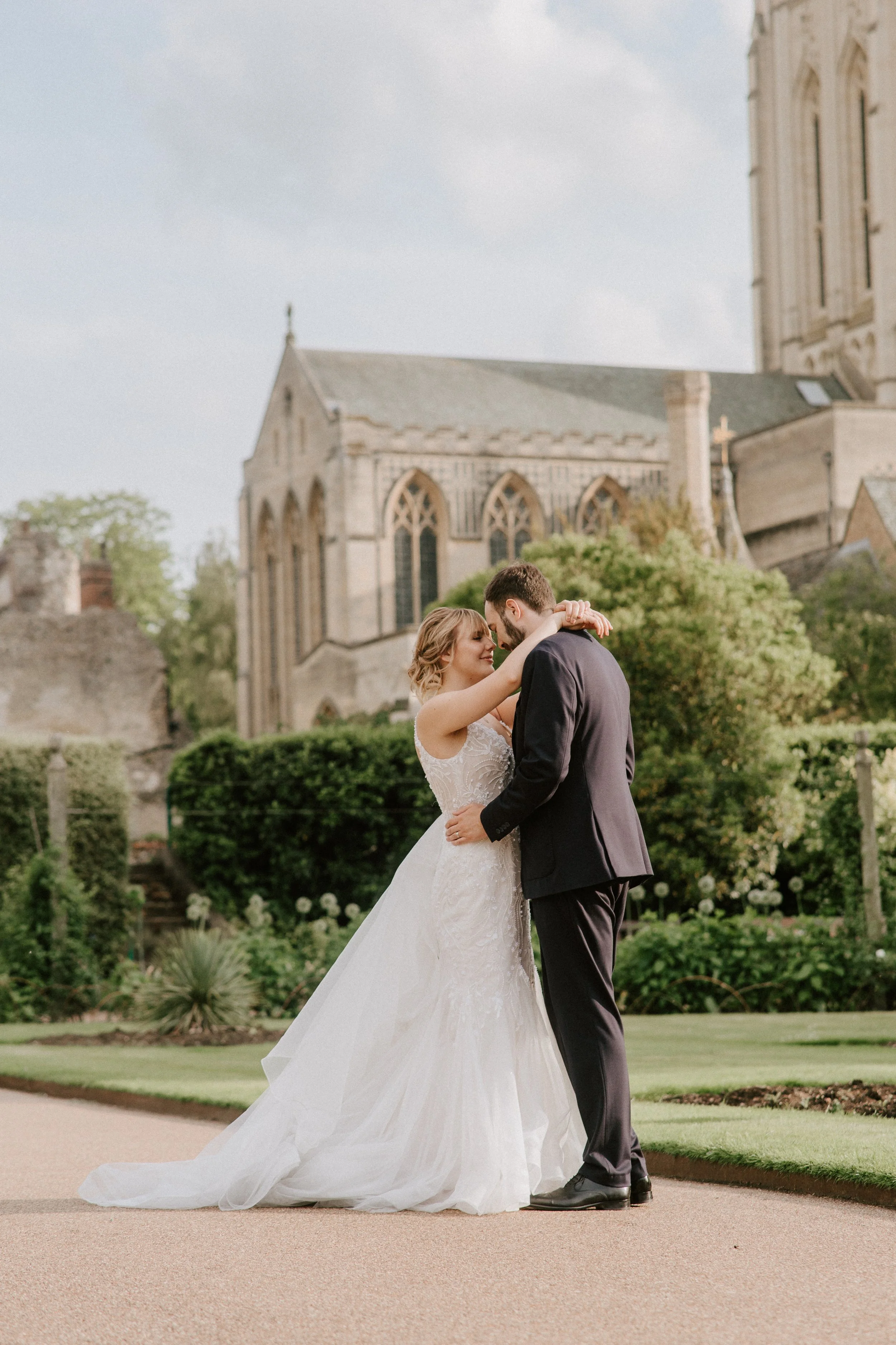 A bride and groom share a romantic dance outdoors in front of a historic church, with greenery and a partly cloudy sky in the background.