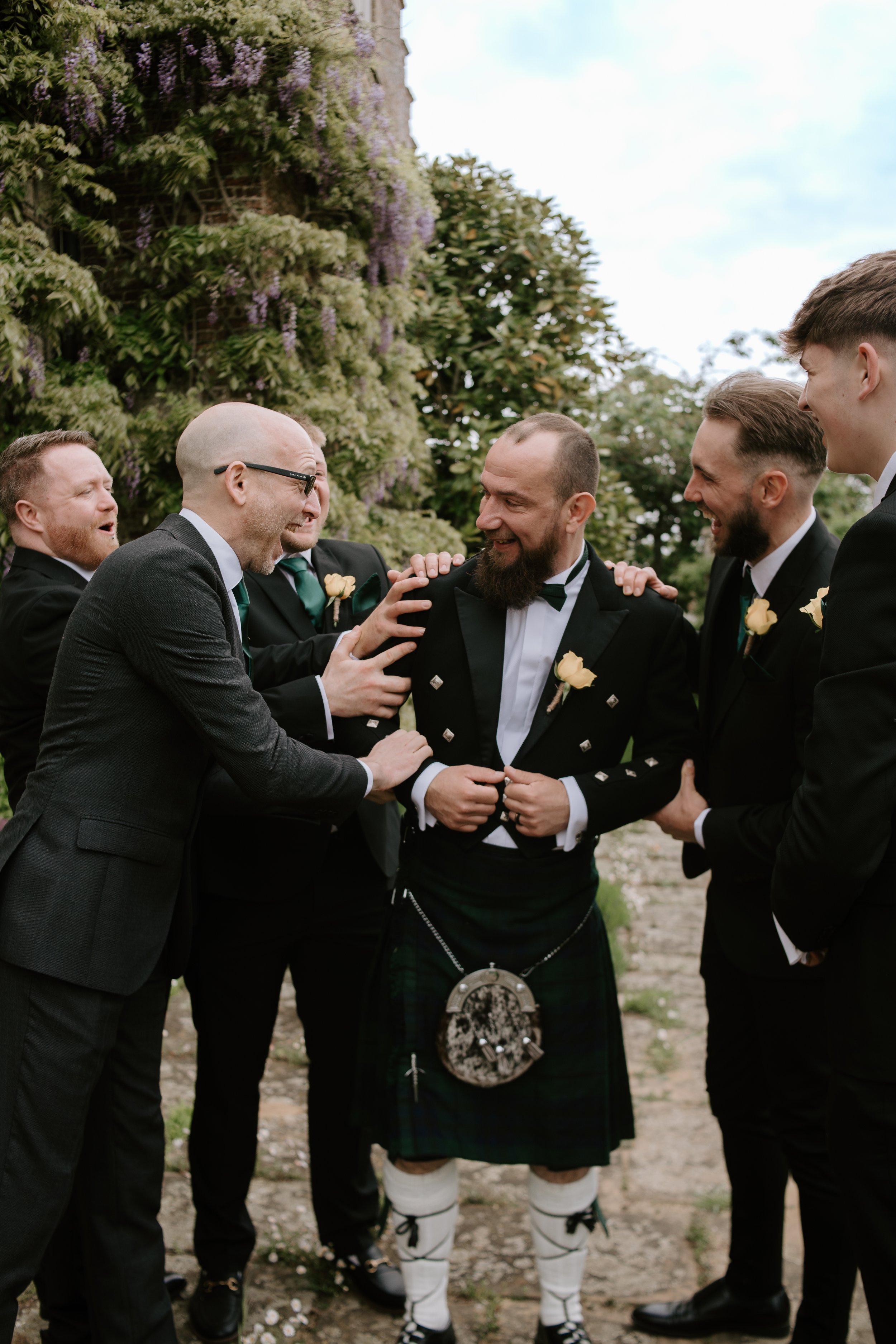 Group of men dressed in formal wear, including a man in traditional Scottish kilt and accessories, smiling and laughing outdoors with lush greenery and purple flowering vines in the background.