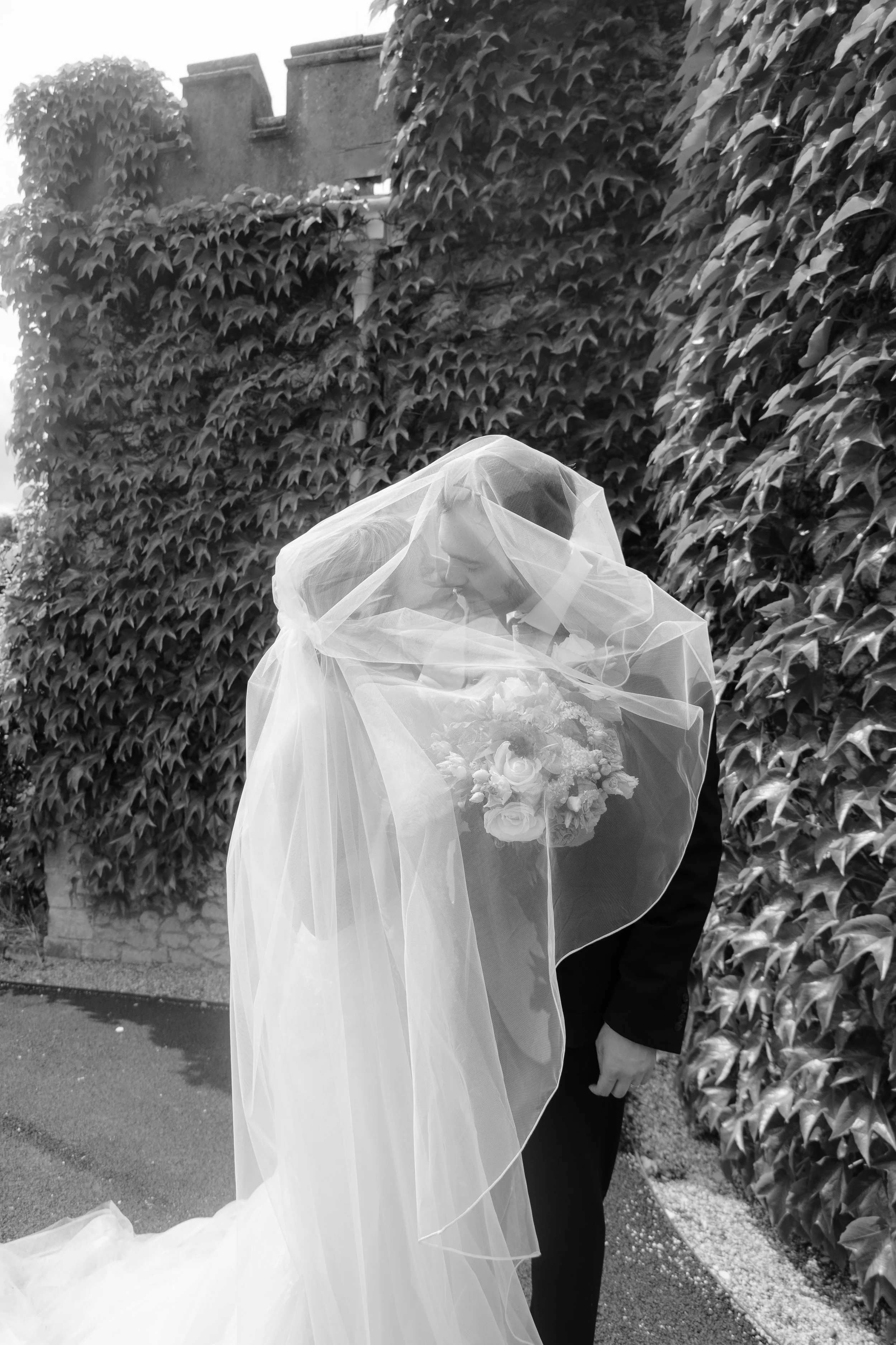 Black and white photo of a bride and groom embracing, with the bride's face partially obscured by a veil and the groom holding a bouquet of flowers. They are standing outdoors near a wall covered with ivy.