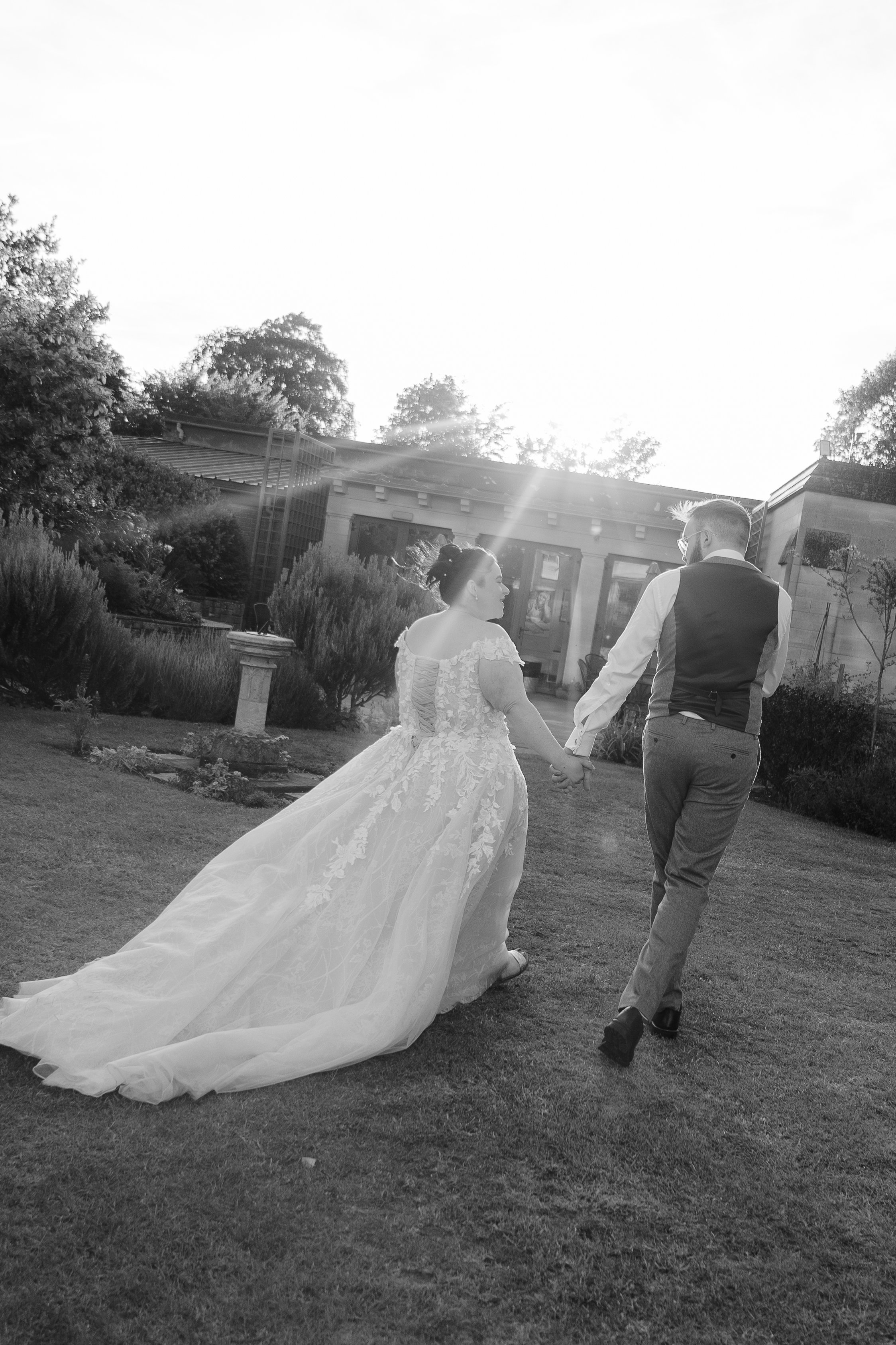 Black and white photo of a bride and groom walking hand in hand on a lawn, with the bride wearing a long flowing wedding dress and the groom in a vest and trousers, backlit by the setting sun.