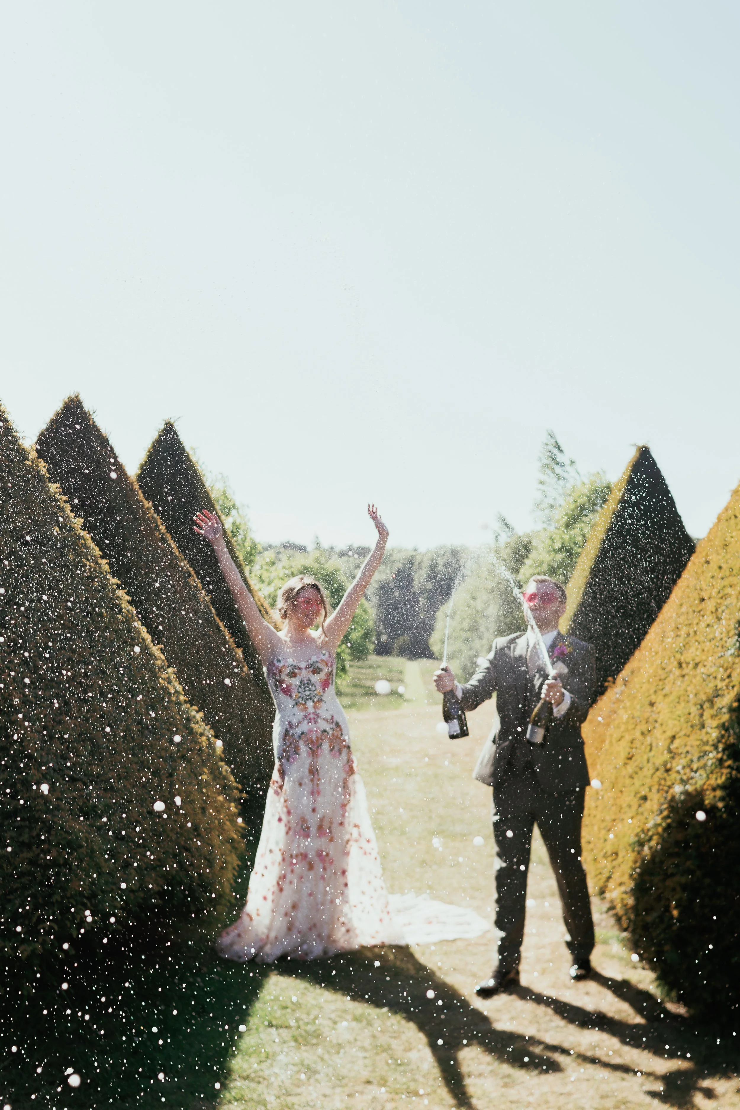A bride and groom celebrating outdoors with champagne bottles, and champagne spraying in the air, surrounded by tall, manicured bushes on a sunny day.