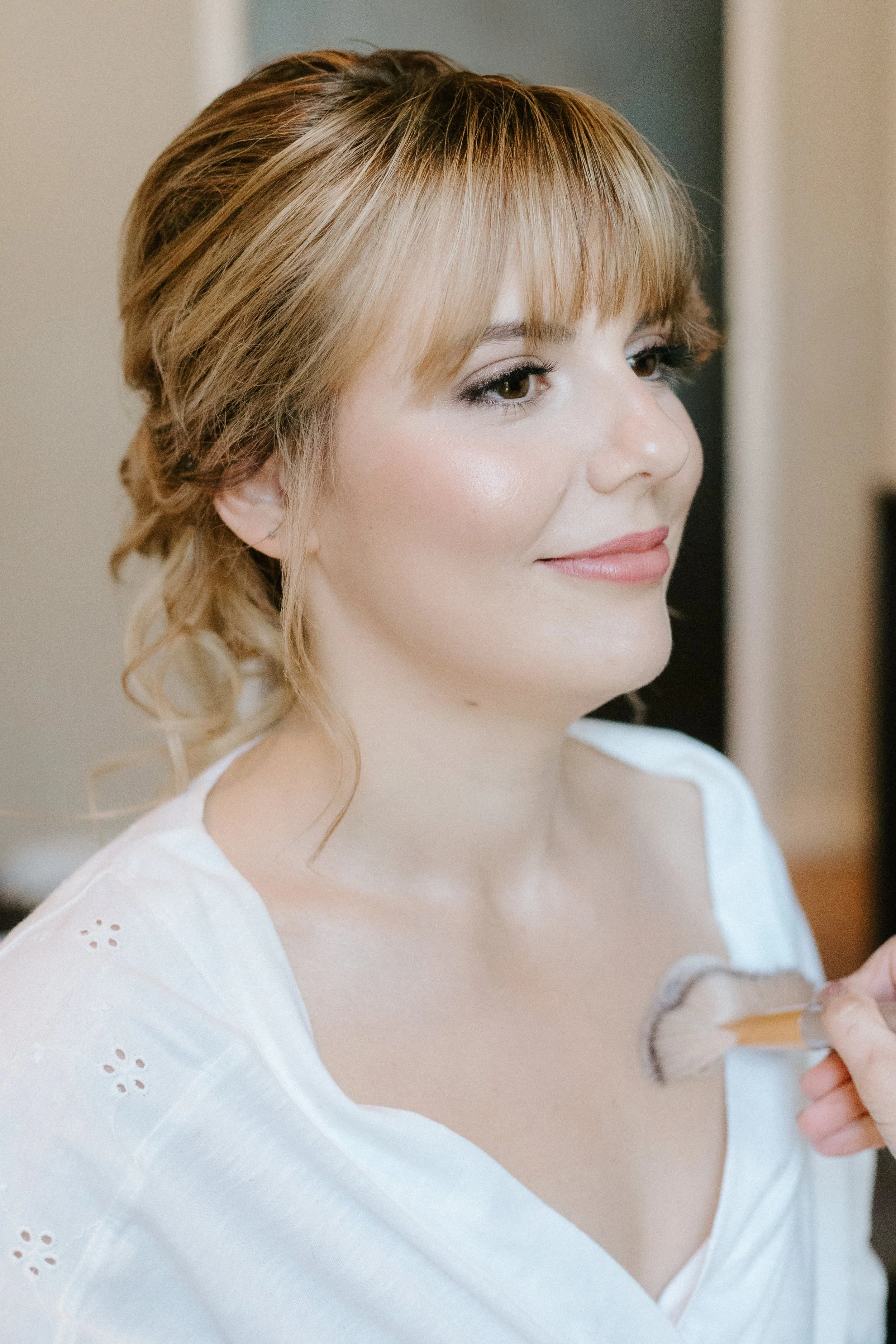 Woman with light brown hair styled in loose curls, receiving makeup application on her chest with a makeup brush.