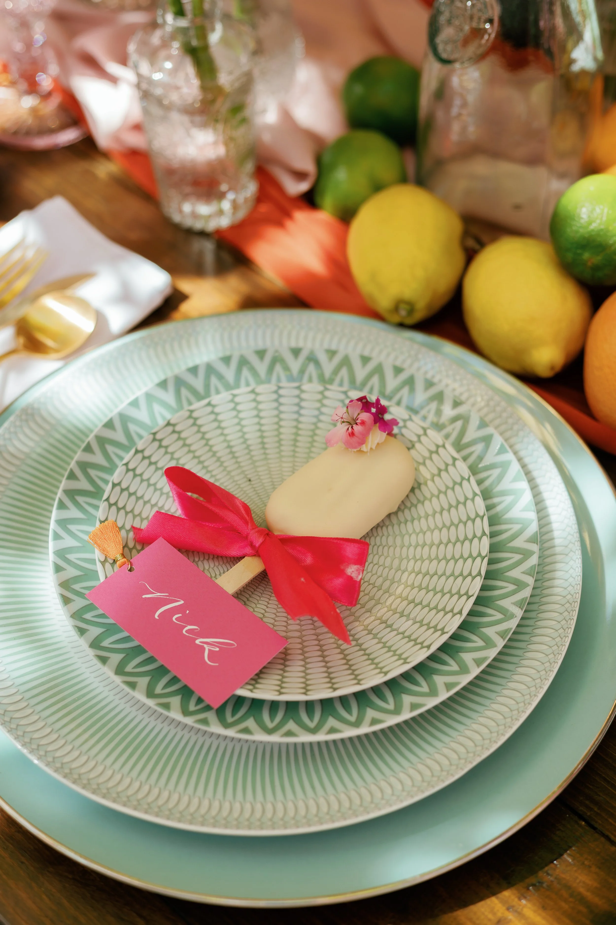 A dessert on a decorative plate with a pink 'Wish' tag, pink flowers, red ribbon, and a white chocolate coating, set on an elegant table with fruit and glassware in the background.