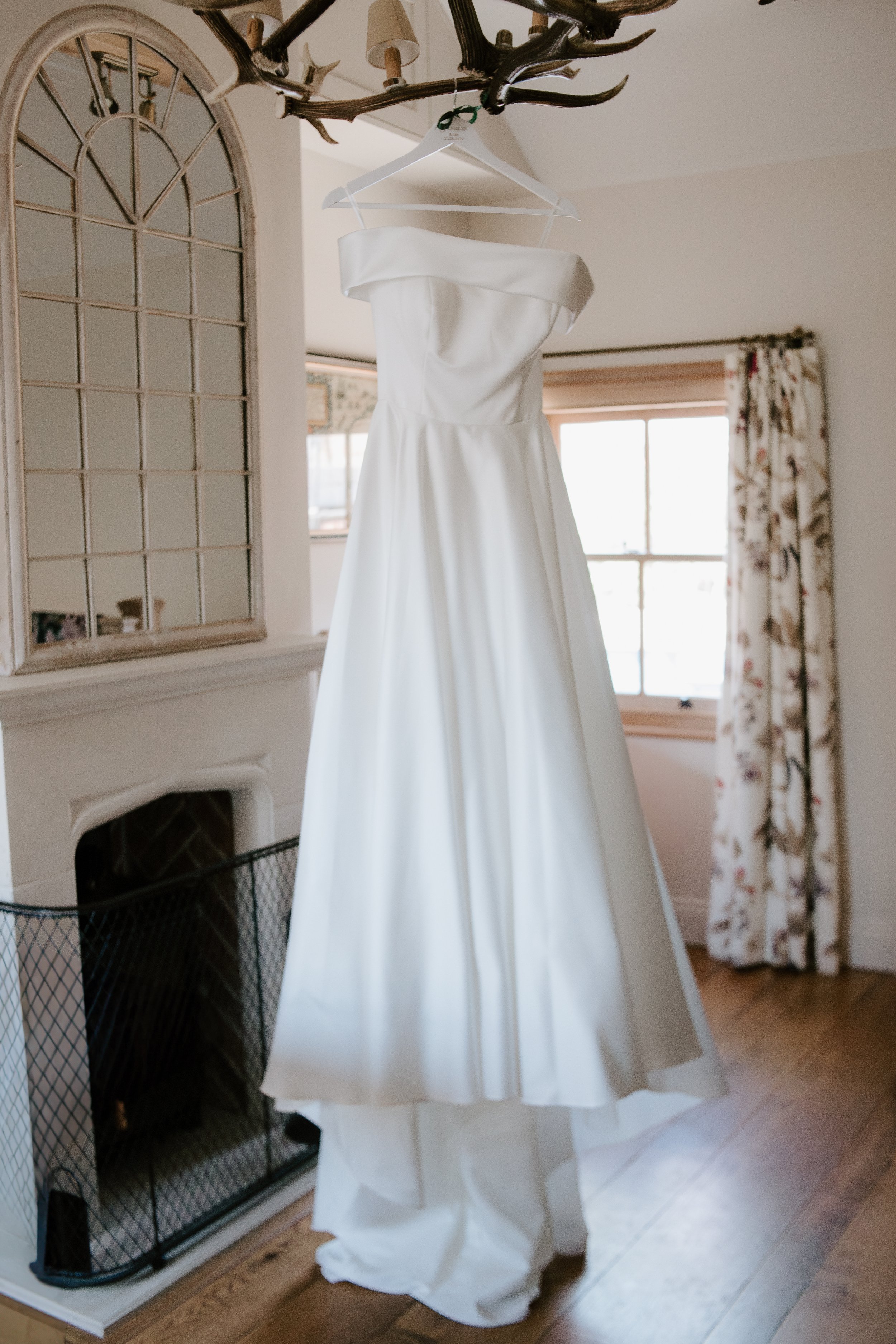 A white wedding dress hanging from a chandelier in a cozy room with a window and floral curtains.