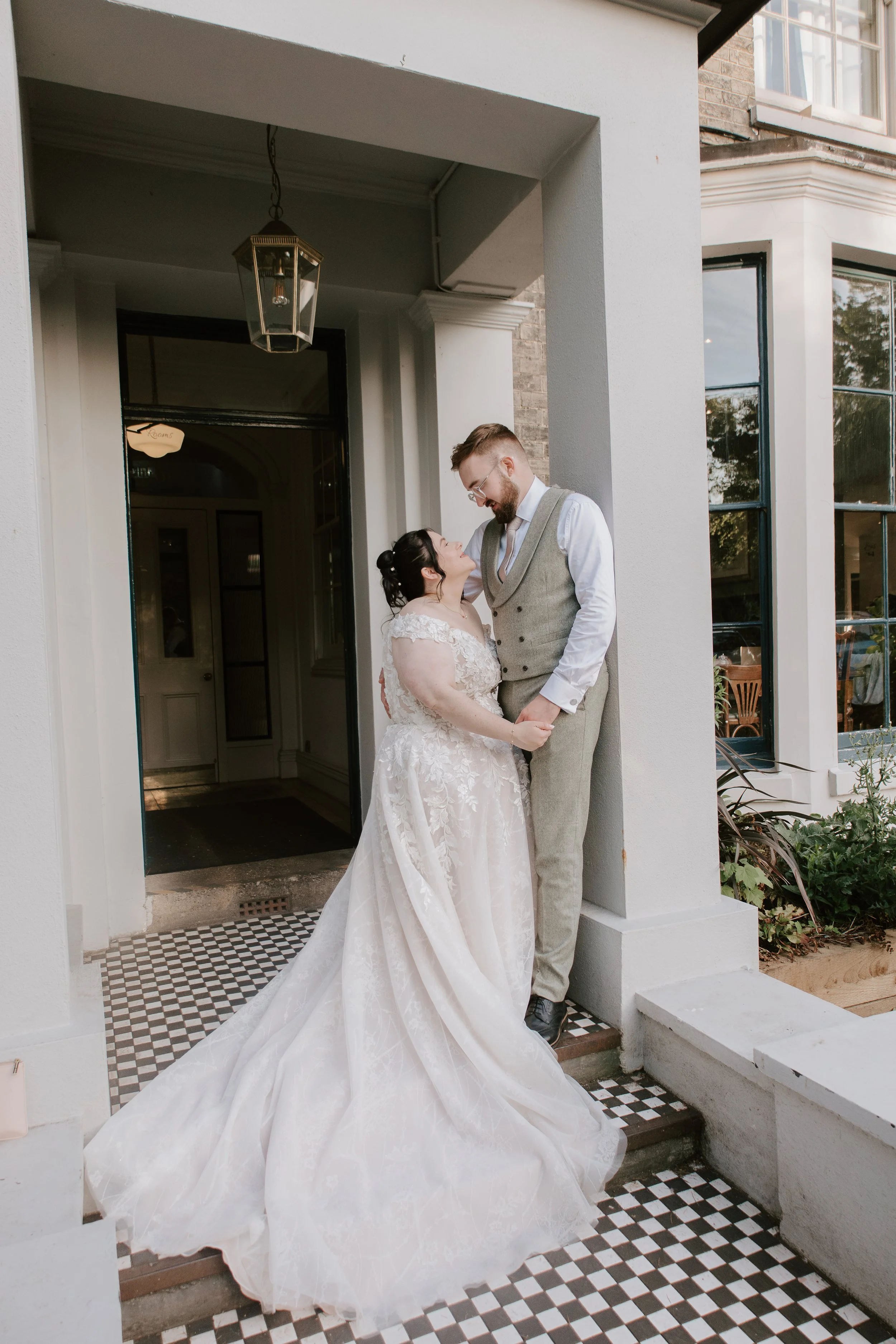 A bride in a lace wedding gown and a groom in a light gray vest and pants share a romantic moment on the front porch of a house, holding hands and gazing into each other's eyes.
