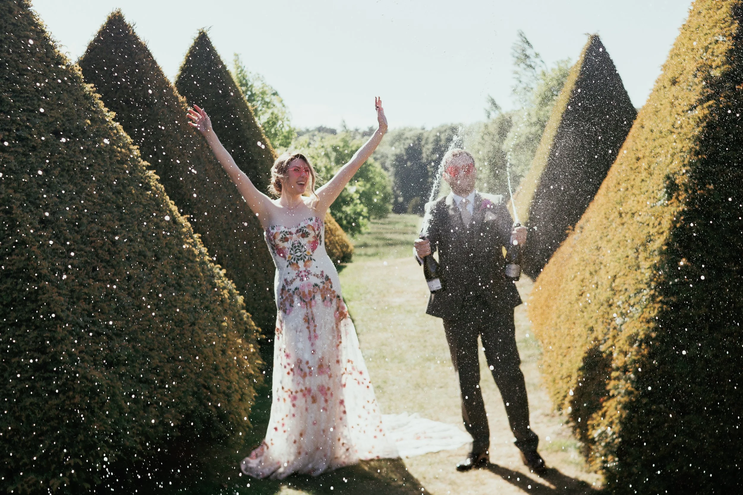 A woman in a floral dress celebrating with her arms raised, standing next to a man in a suit with champagne bottles spraying champagne between them in a garden with tall bushes.