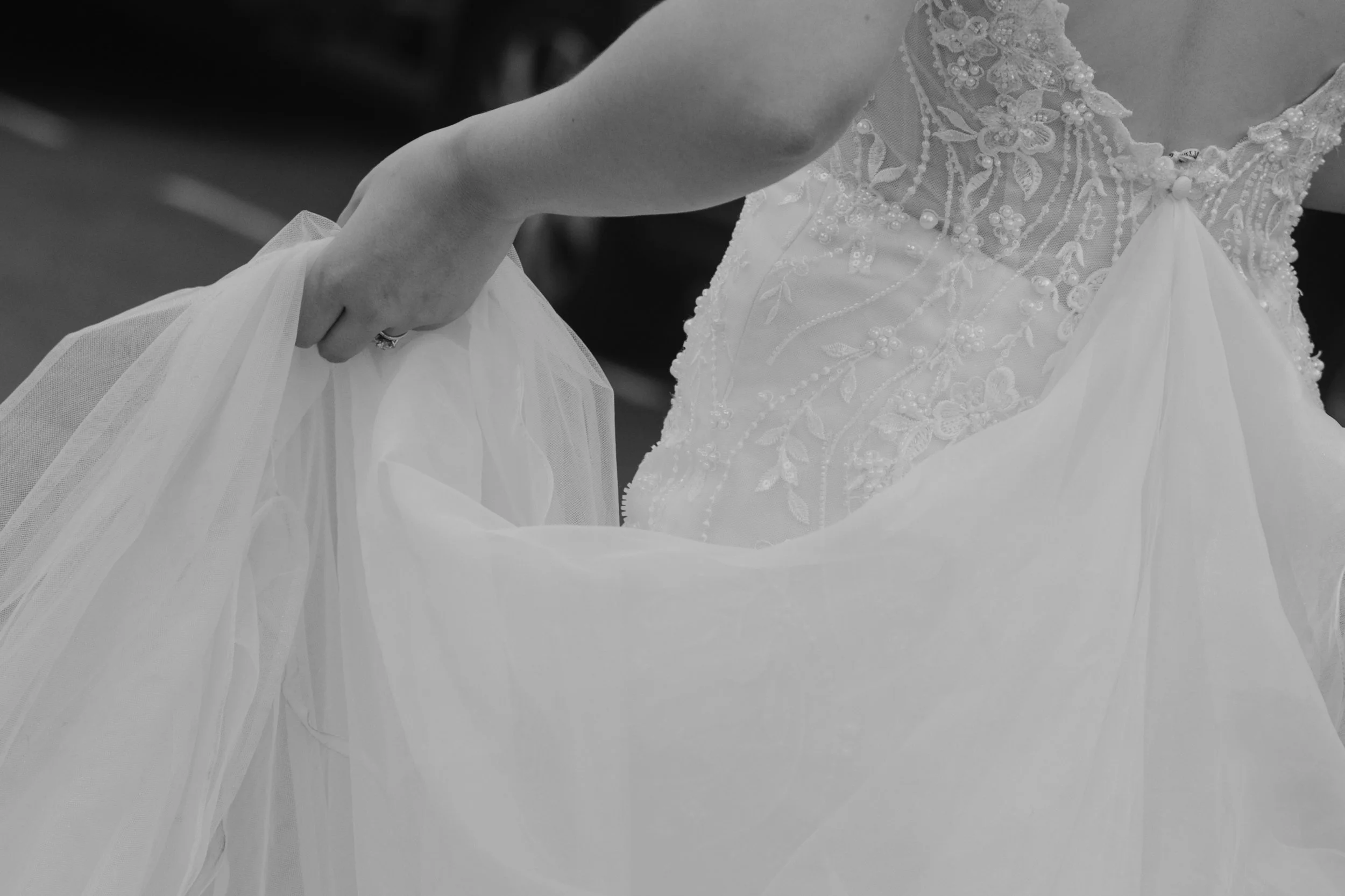 A bride holding the skirt of her wedding dress, featuring intricate lace and bead embroidery, with a focus on her hand and dress fabric.