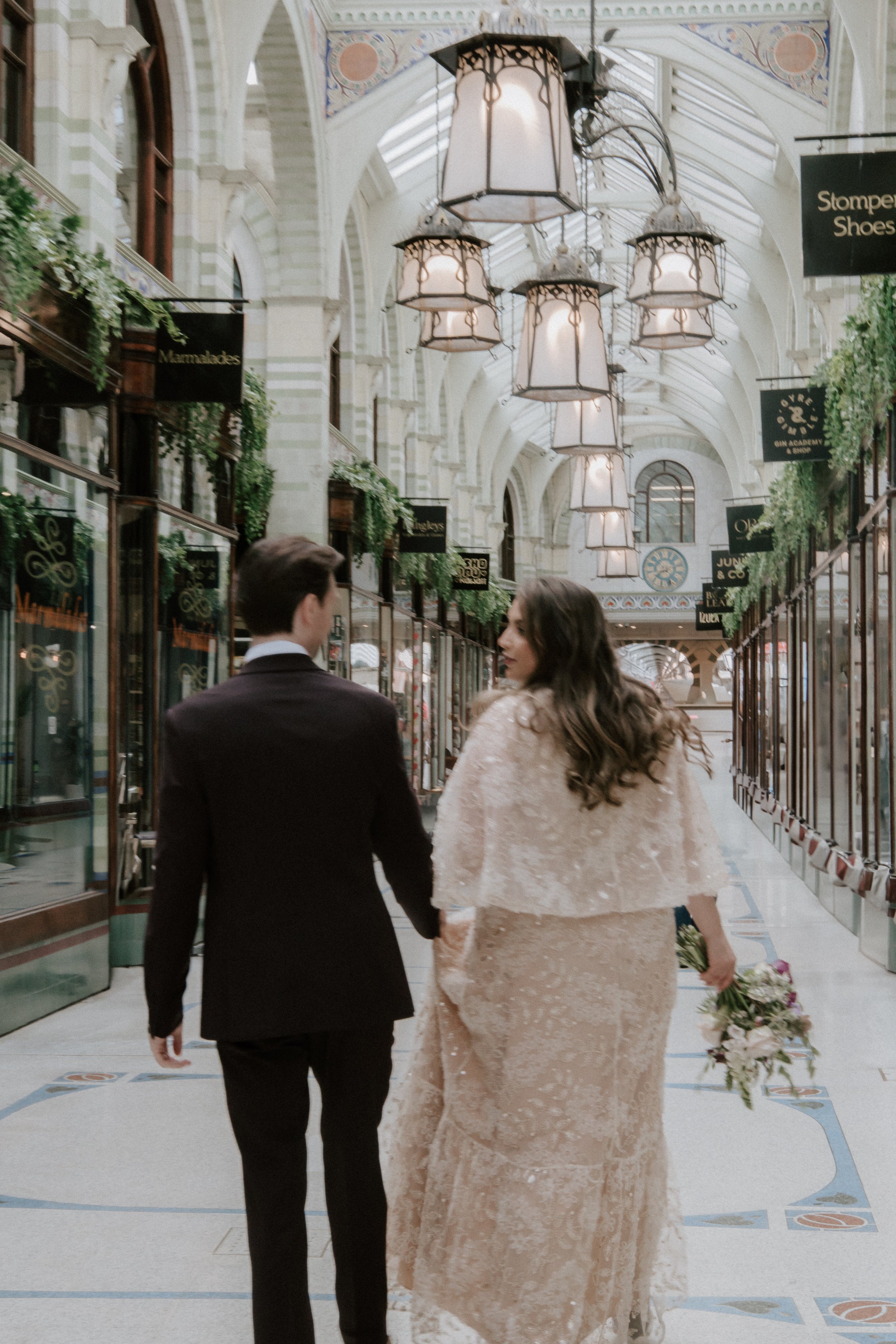 A man and a woman walking hand in hand inside a grand, historic shopping arcade with ornate architecture, hanging lamps, and storefronts on either side. The woman is holding a bouquet of flowers.