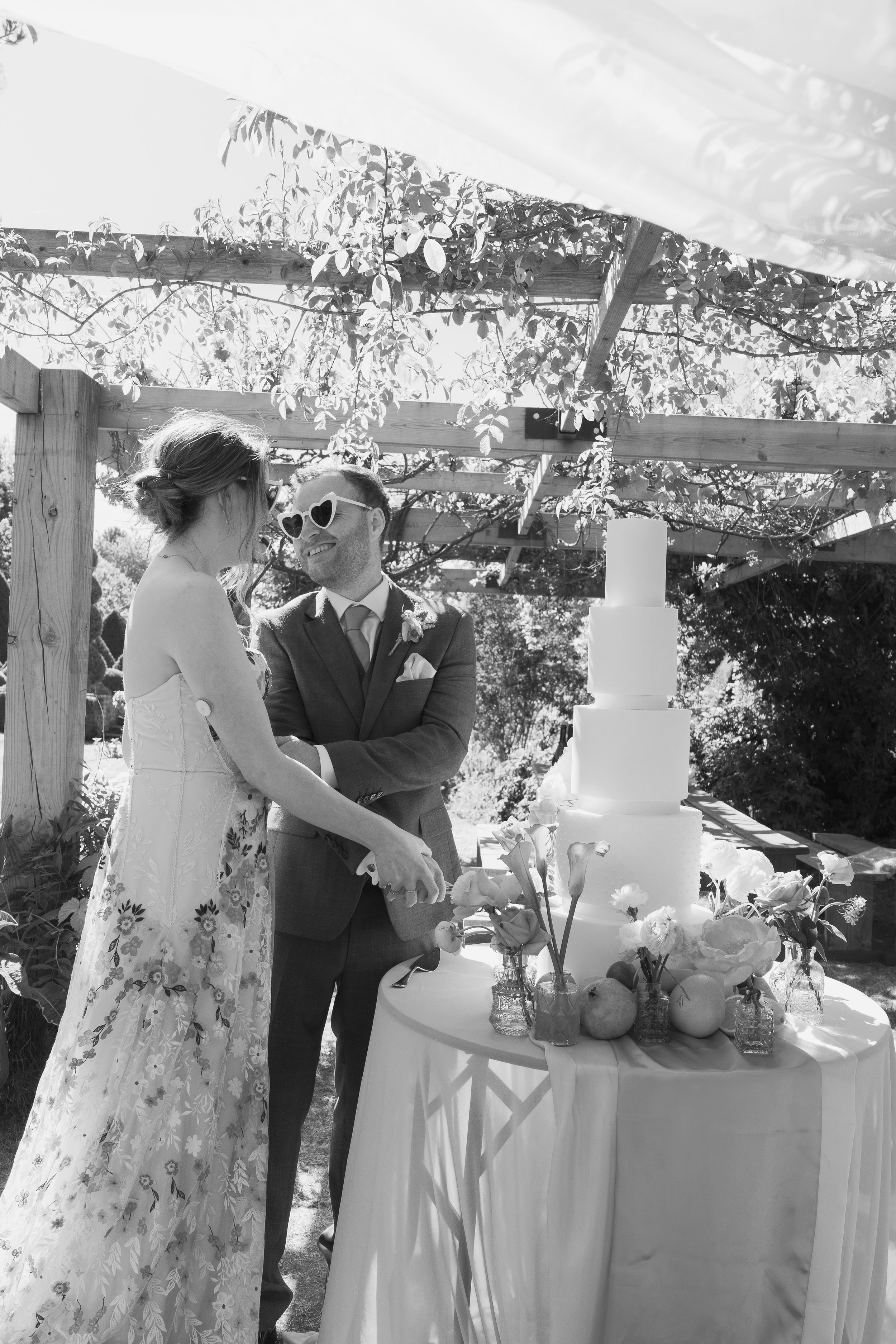 Black and white photo of a bride and groom at their wedding reception, cutting a wedding cake together outdoors under a pergola, with flowers and fruits on the table.