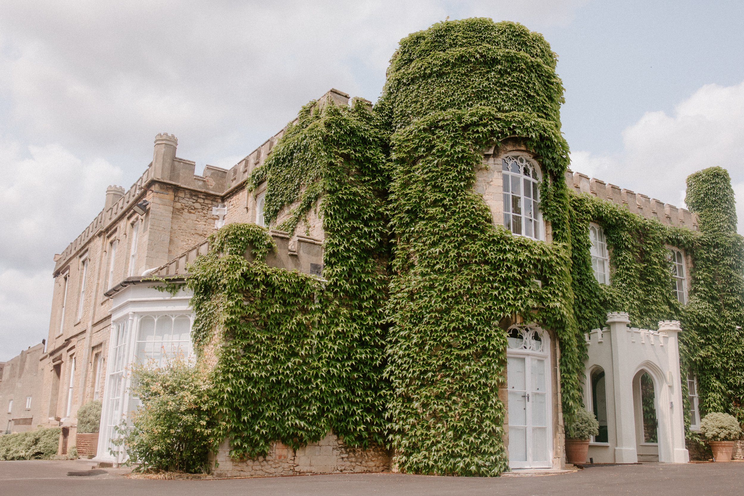 A large stone building covered in green ivy with white-framed windows and a white entrance door, set against a cloudy sky.