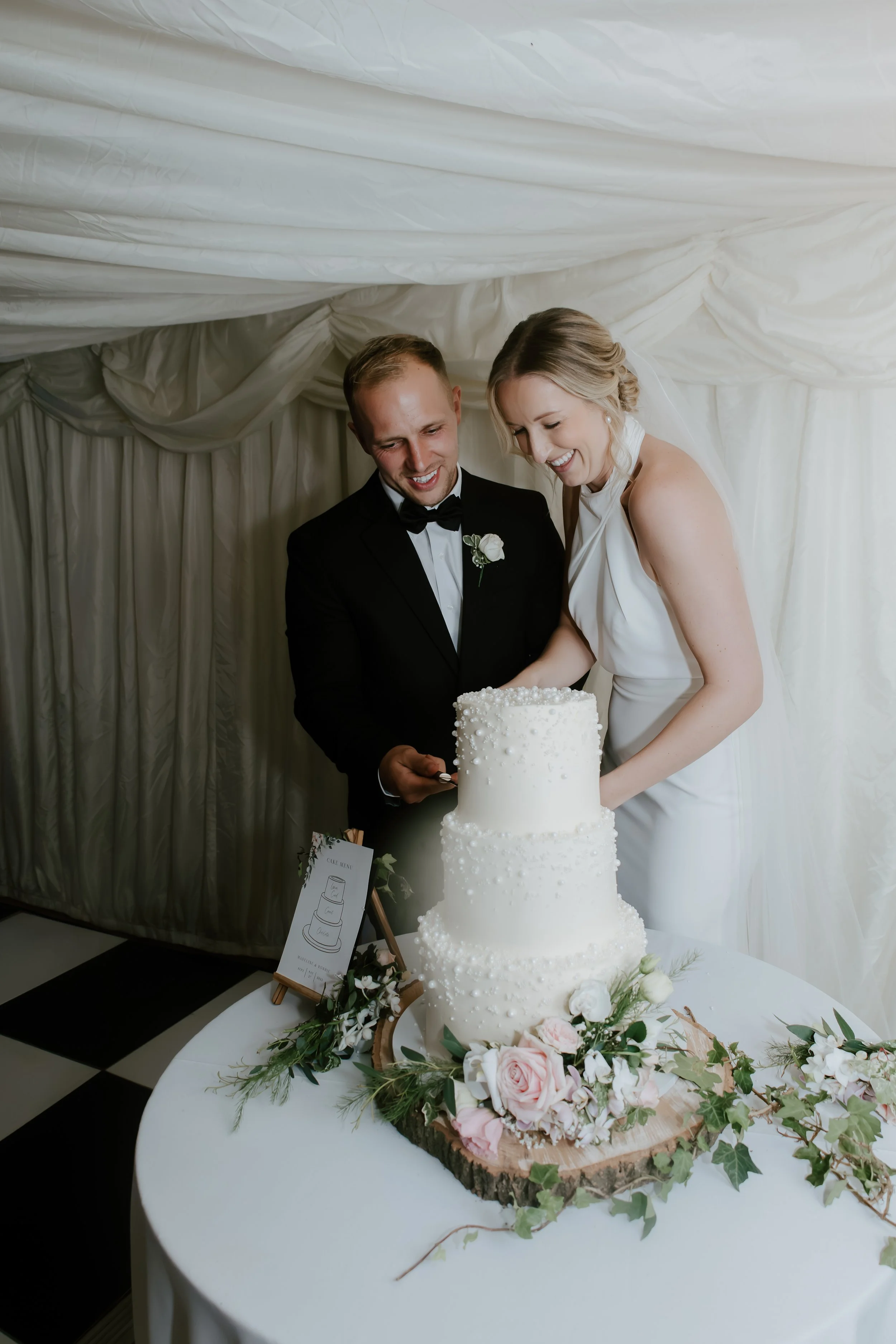 Elegant and classic wedding photographer Suffolk and Norfolk – bride and groom cutting wedding cake decorated with white pearls surrounded by pink and white flowers.