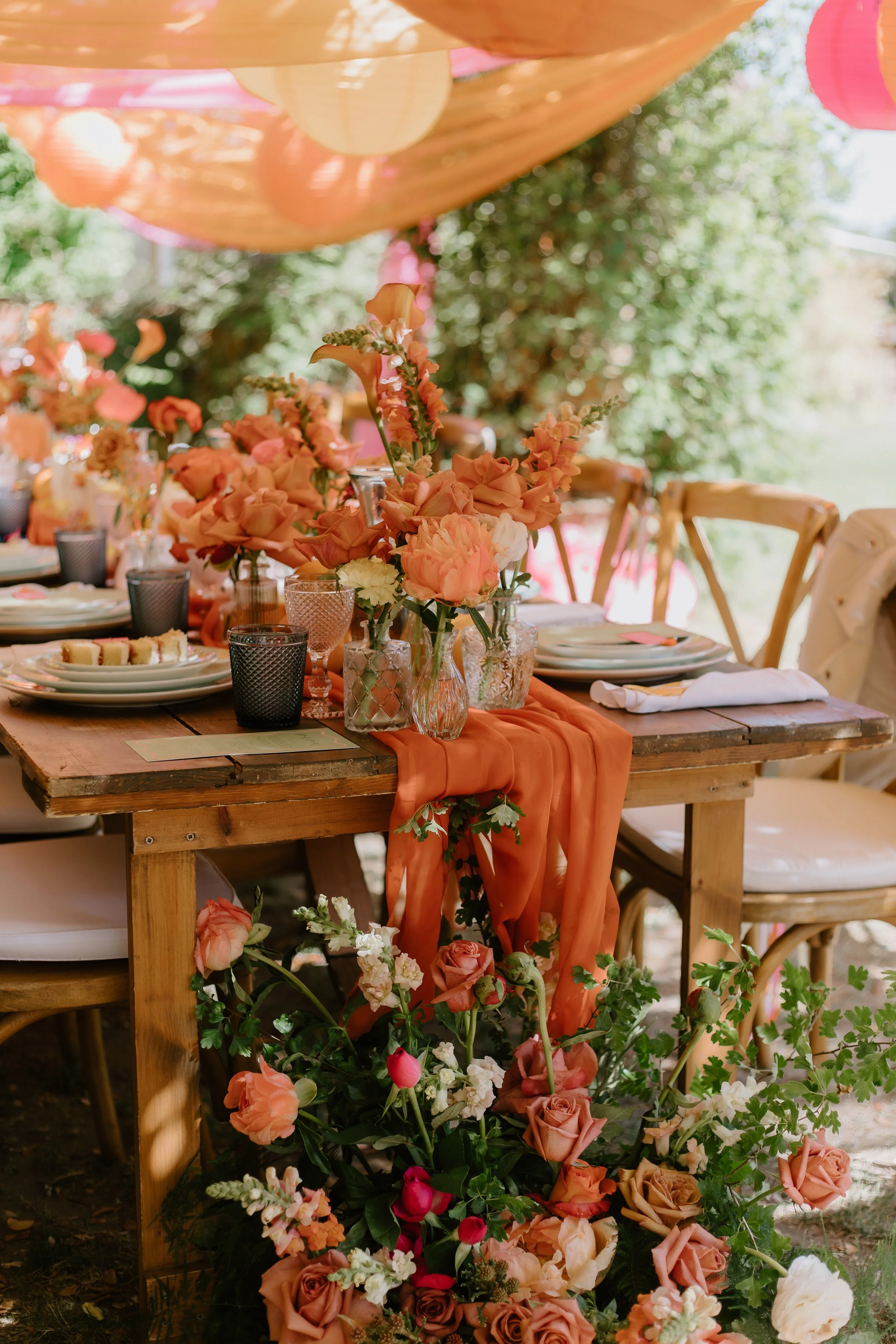 A beautifully decorated outdoor dining table with peach and pink flowers, orange fabric, and paper lanterns hanging above.