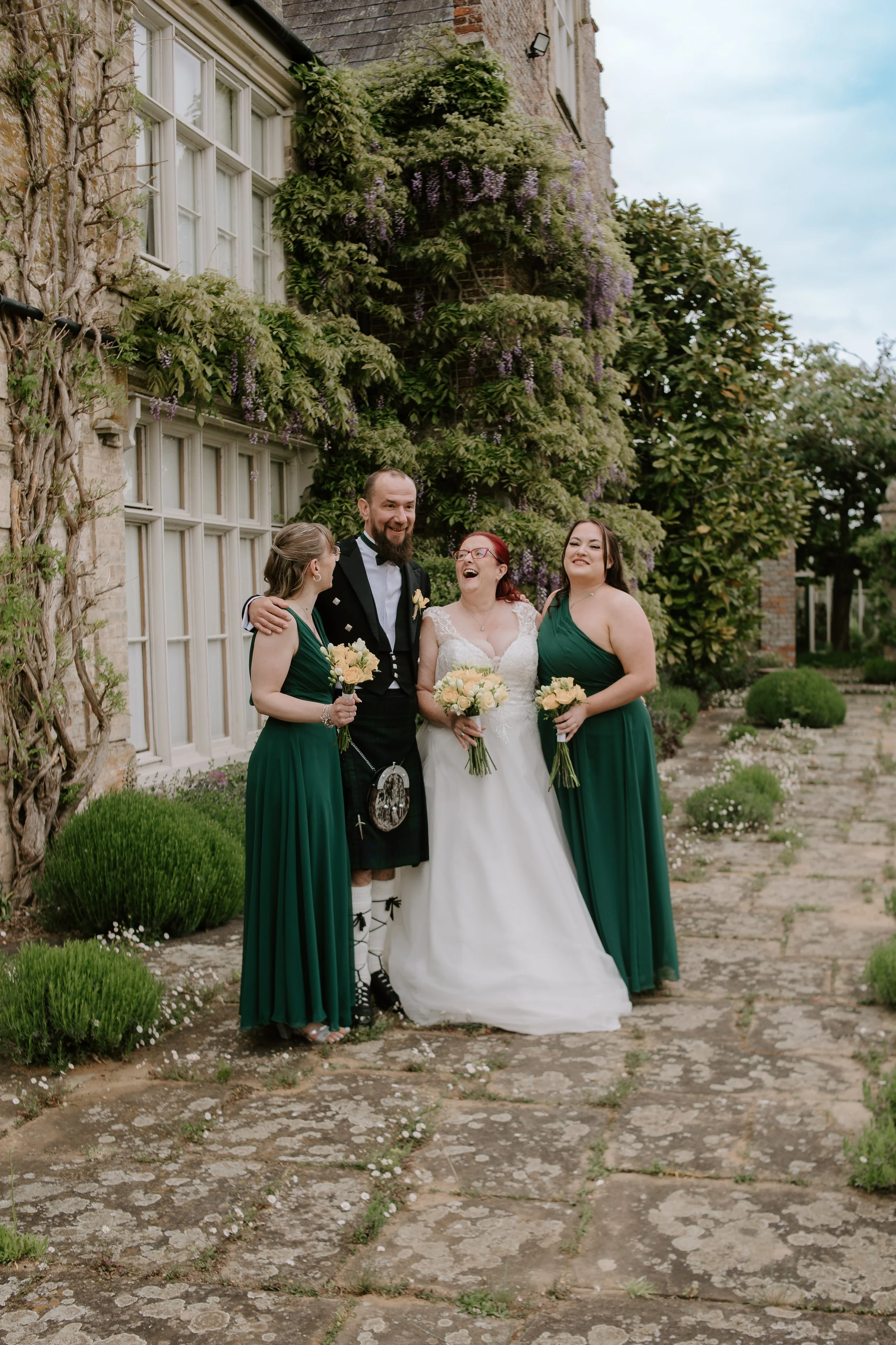 A bride in a white wedding gown standing between two bridesmaids in green dresses and a groom dressed in traditional Scottish attire, all smiling and holding bouquets, outside a building with lush greenery and flowers.