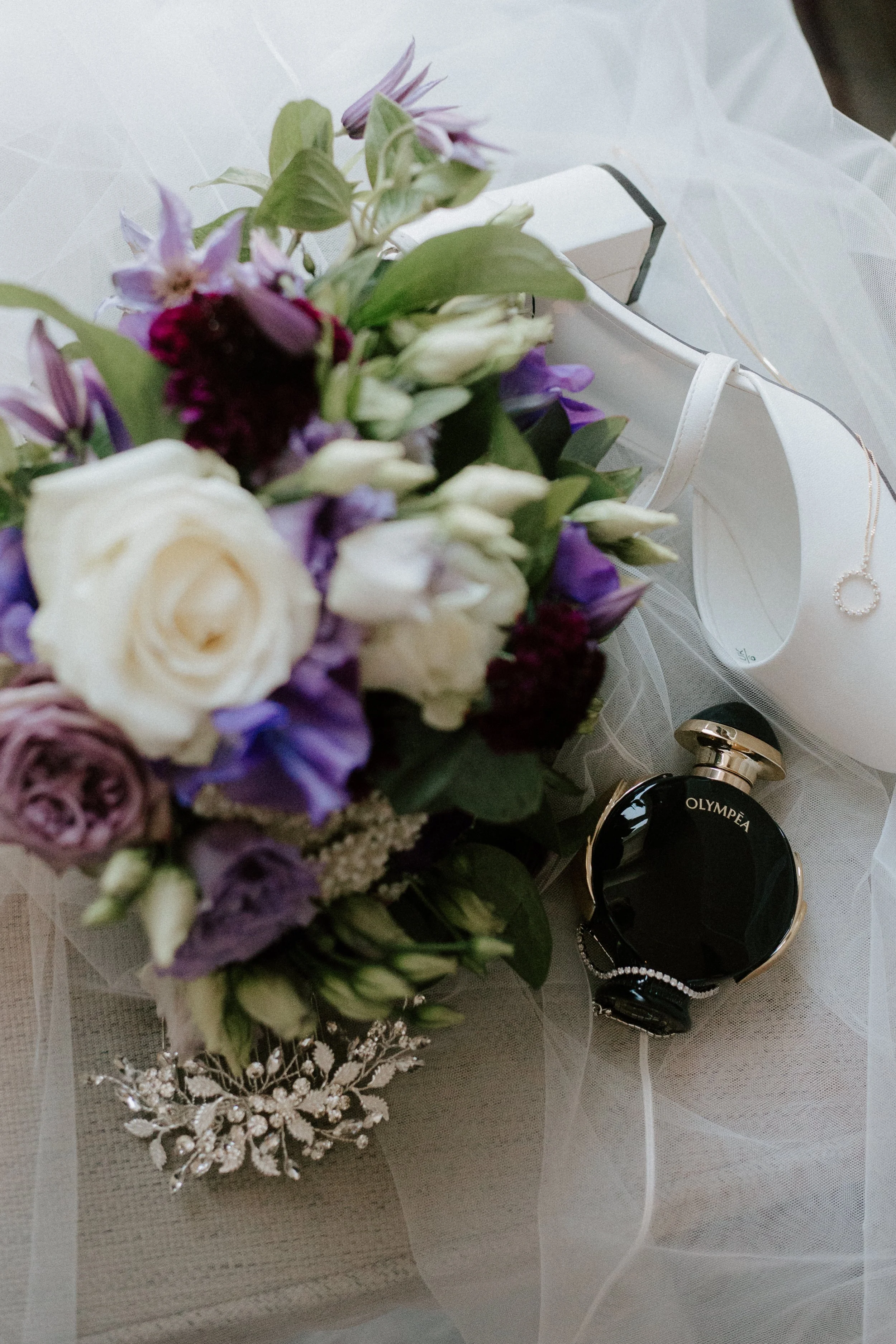 A bouquet of flowers, a bottle of black perfume labeled 'OLYMPEA,' a white high-heeled shoe, a diamond bracelet, and a silver hairpiece with crystals resting on a light-colored surface with tulle fabric.