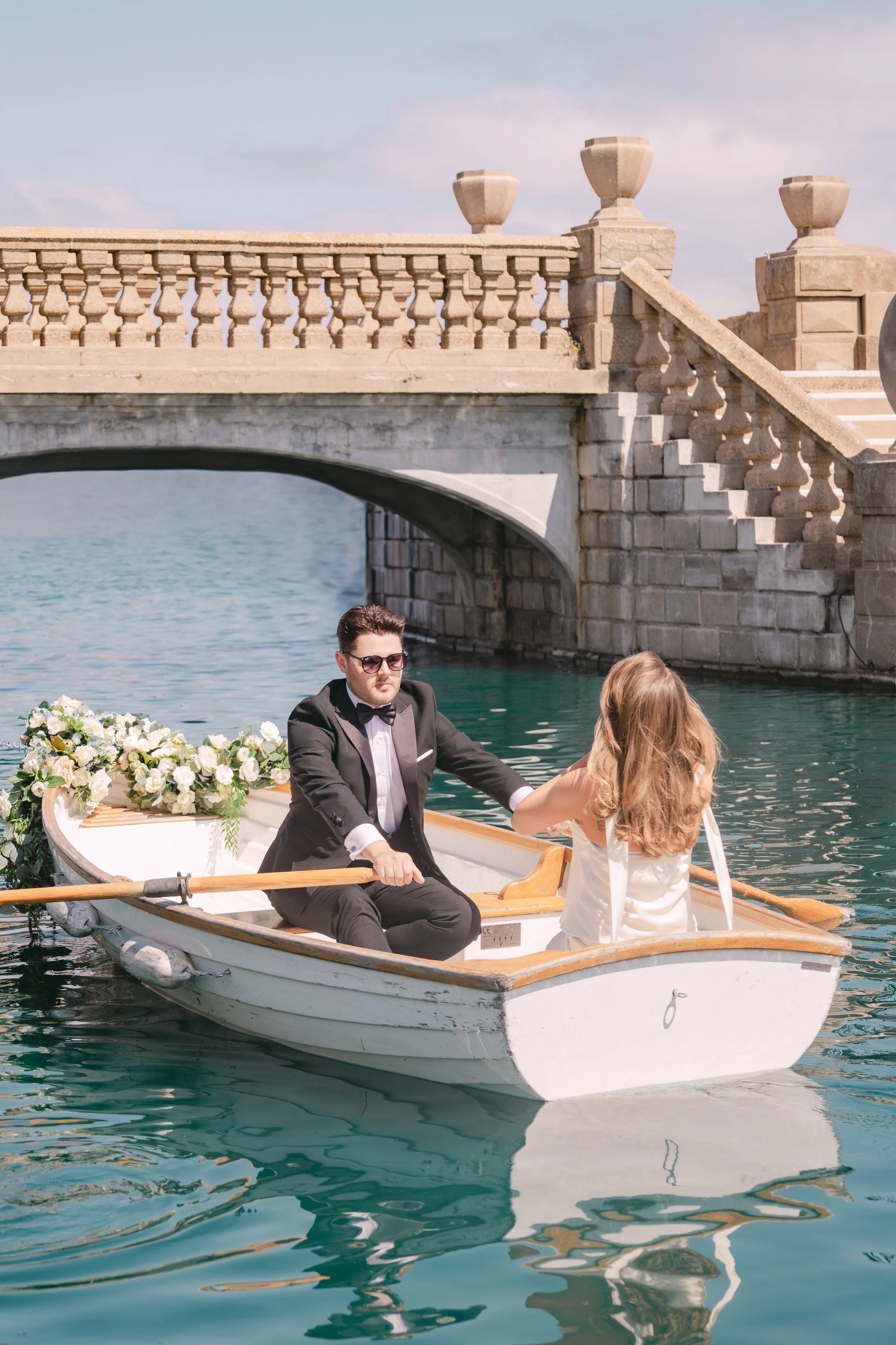 A man in a tuxedo rowing a small boat with a woman seated in the boat, near a stone bridge over water.