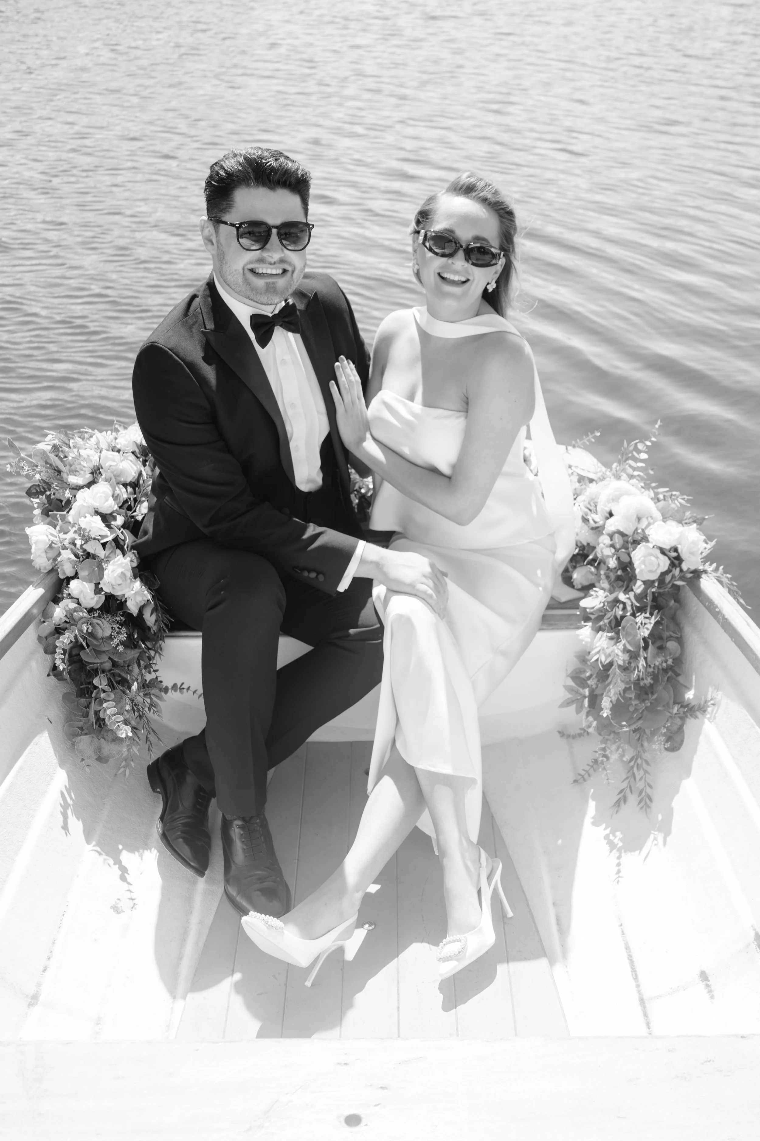 Black and white photo of a wedding couple sitting in a boat on a lake, smiling, with flowers on the boat's side, the bride in a white dress and heels, the groom in a tuxedo and sunglasses.