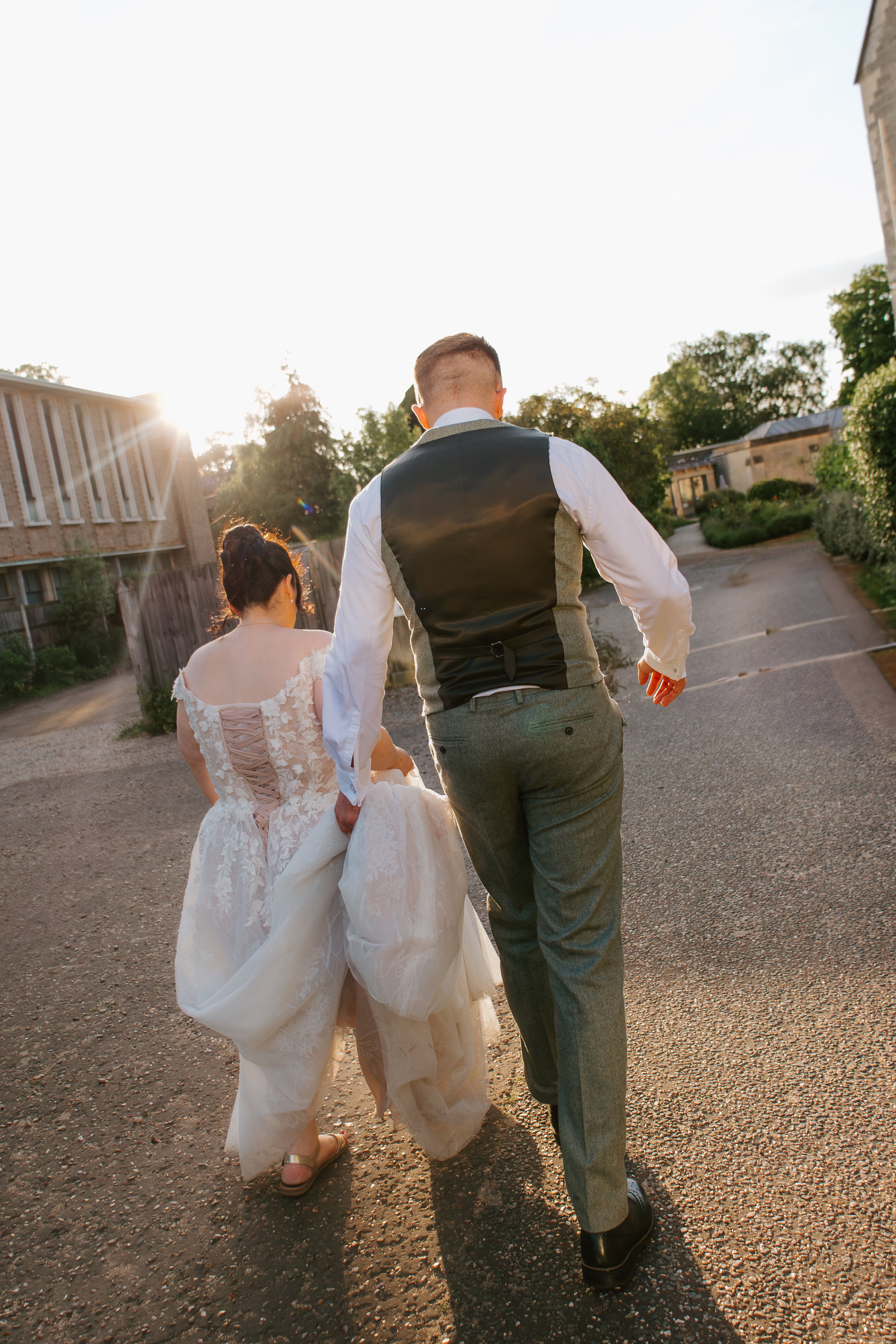 A couple in wedding attire walking outdoors at sunset, with the bride holding her gown and the groom in a suit vest.