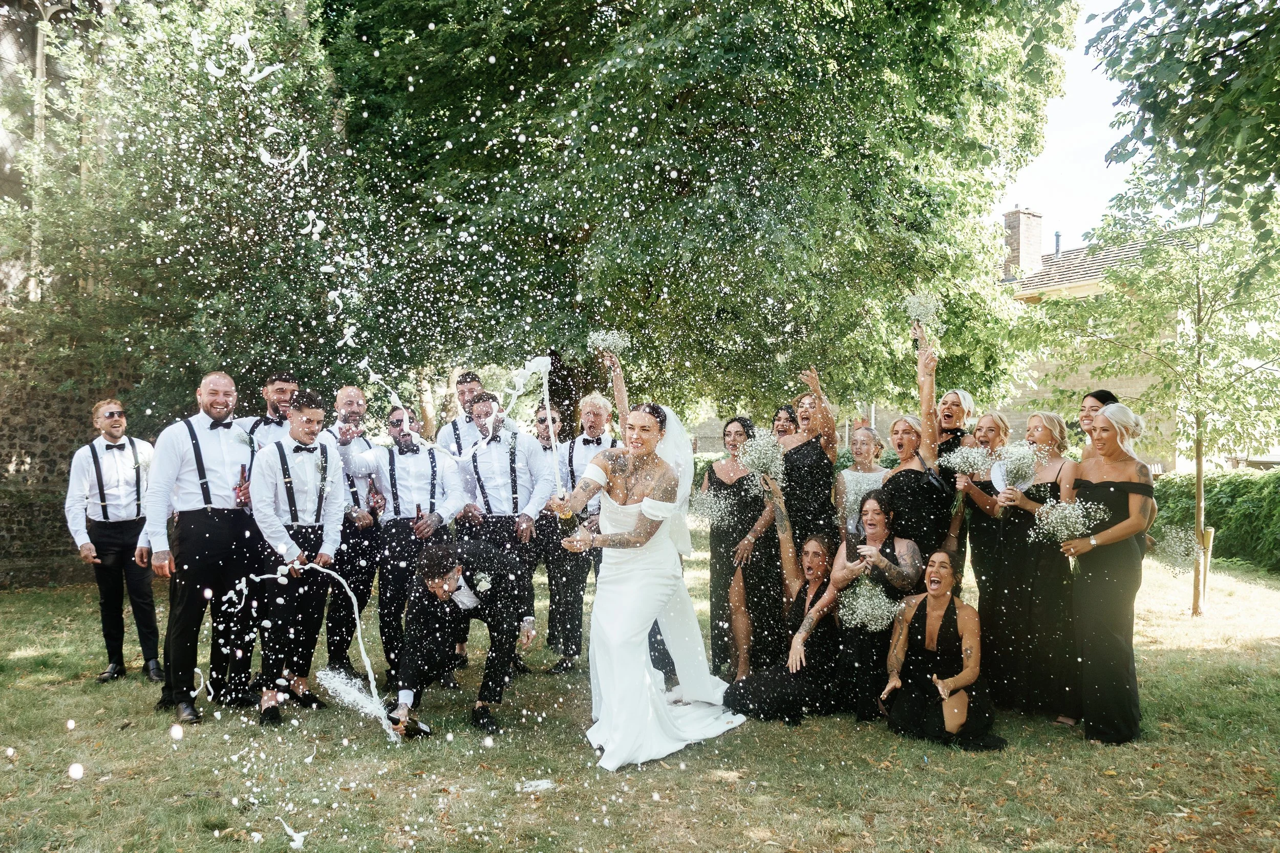 Epic champagne spray moment with full bridal party during summer wedding in Suffolk – candid and fun group photo by UK wedding photographer.