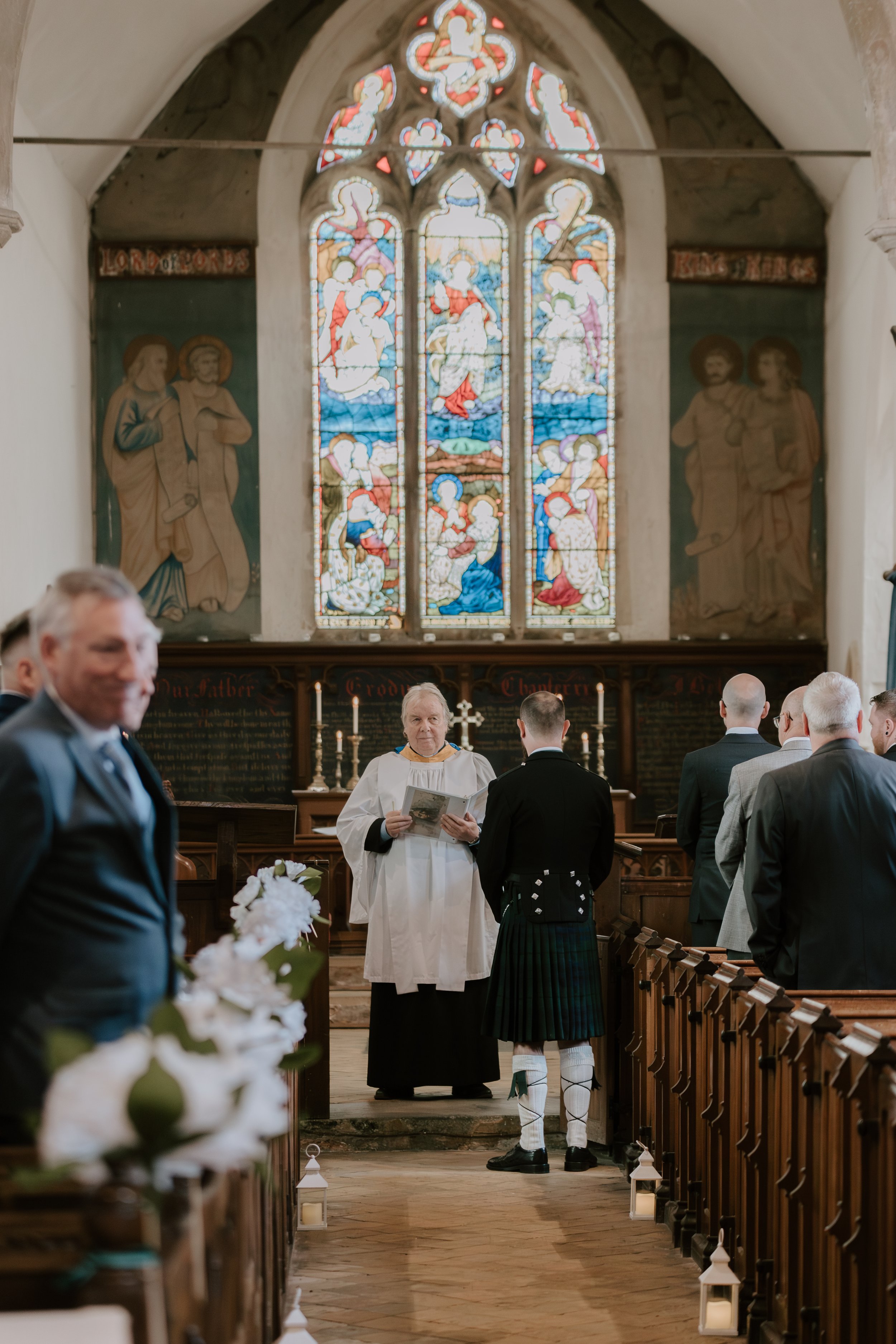 A wedding ceremony taking place inside a church with stained glass windows depicting religious scenes, a minister in white robes, and a groom dressed in traditional Scottish kilt, surrounded by guests.