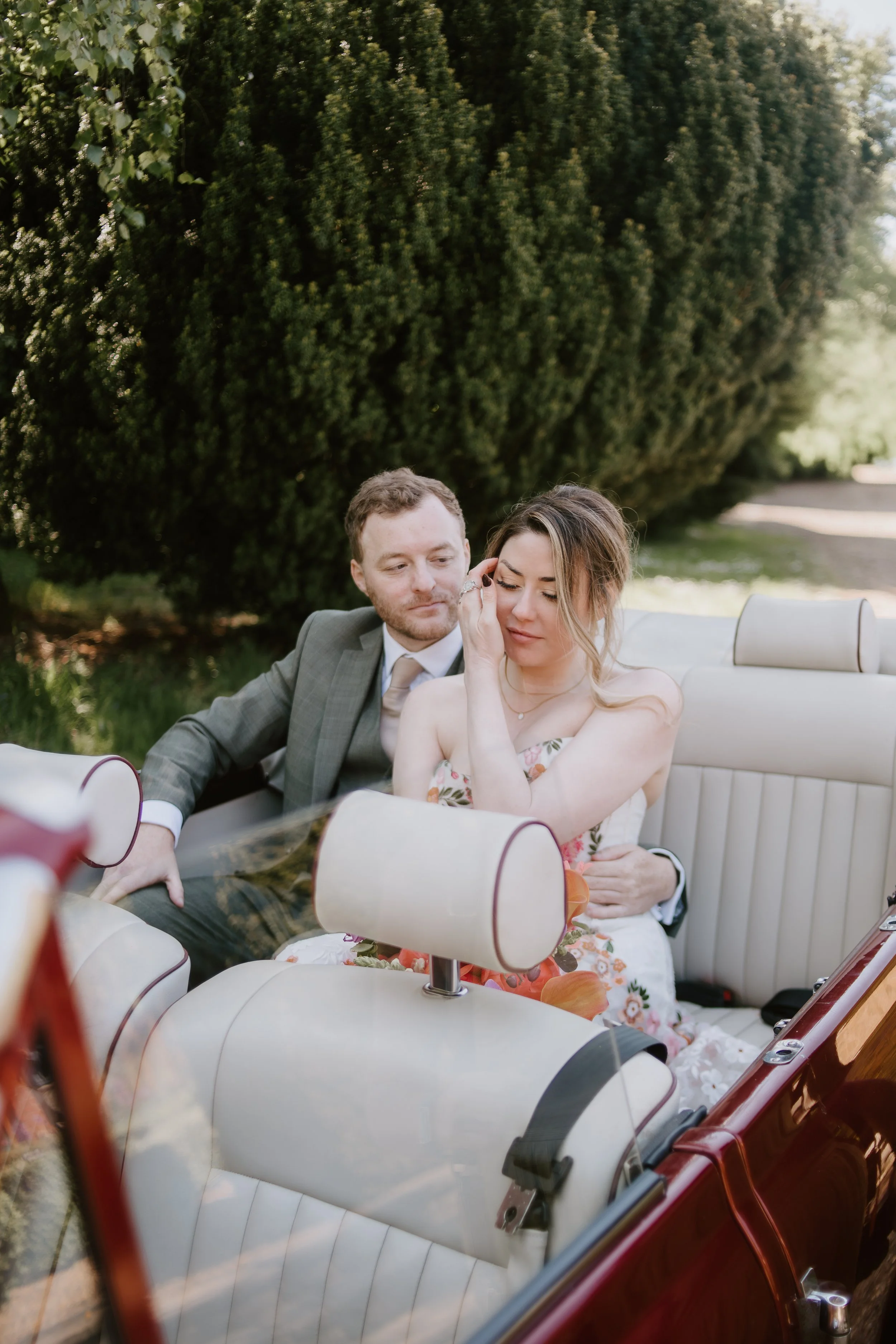 A bride and groom sitting in a vintage car outdoors, with the groom wearing a gray suit and the bride wearing a floral dress, surrounded by greenery.