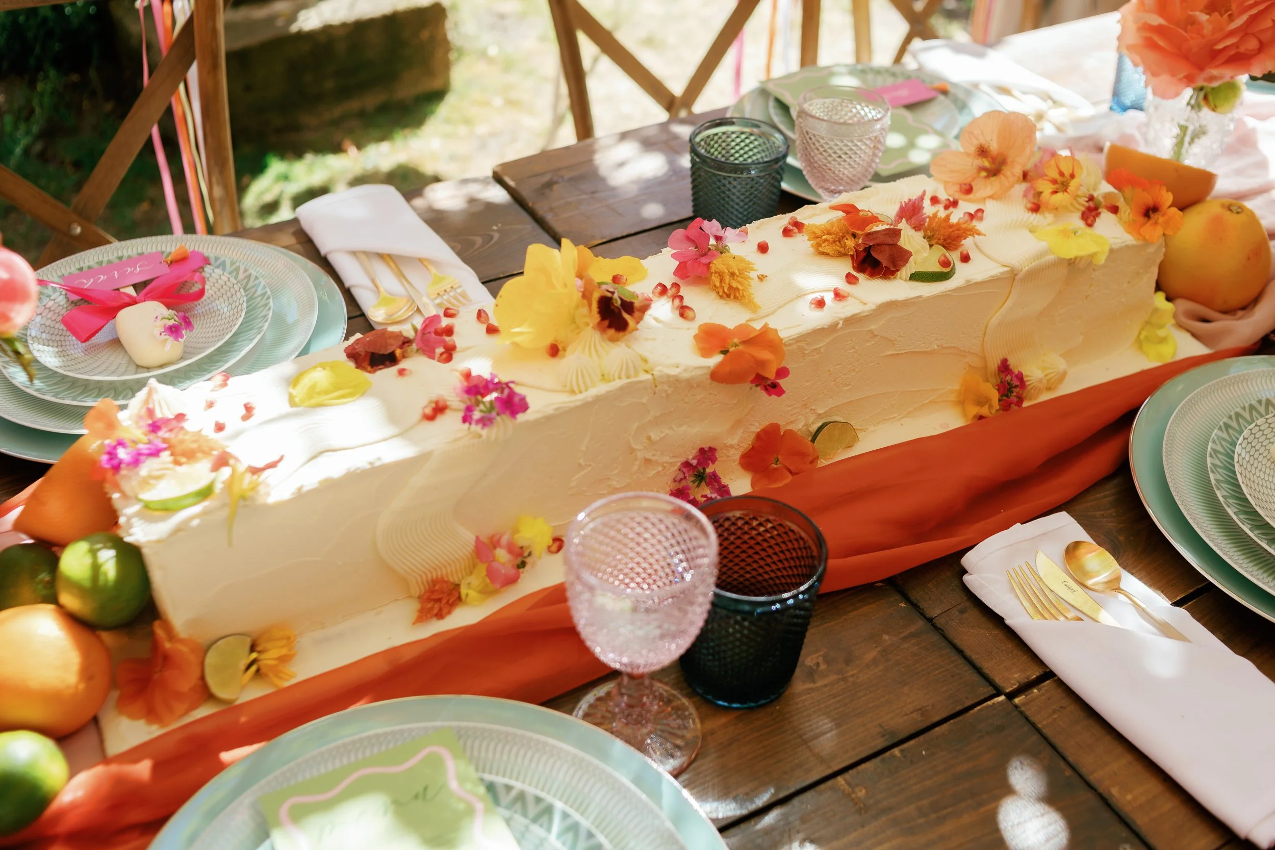 A long white cake decorated with colorful edible flowers on a wooden table, surrounded by tableware including plates, glasses, napkins, and utensils, set outdoors under sunlight.