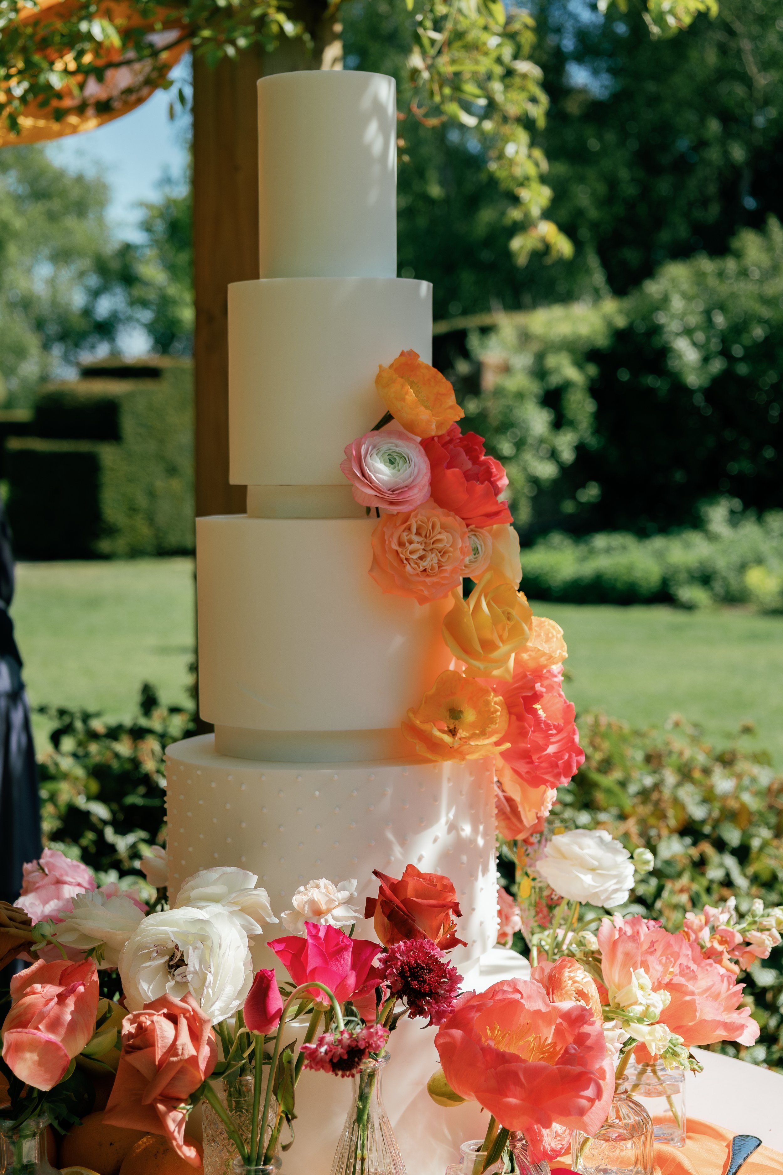 A tall, multi-tiered white wedding cake decorated with fresh pink, orange, and red flowers, surrounded by additional floral arrangements in small vases at an outdoor wedding reception.