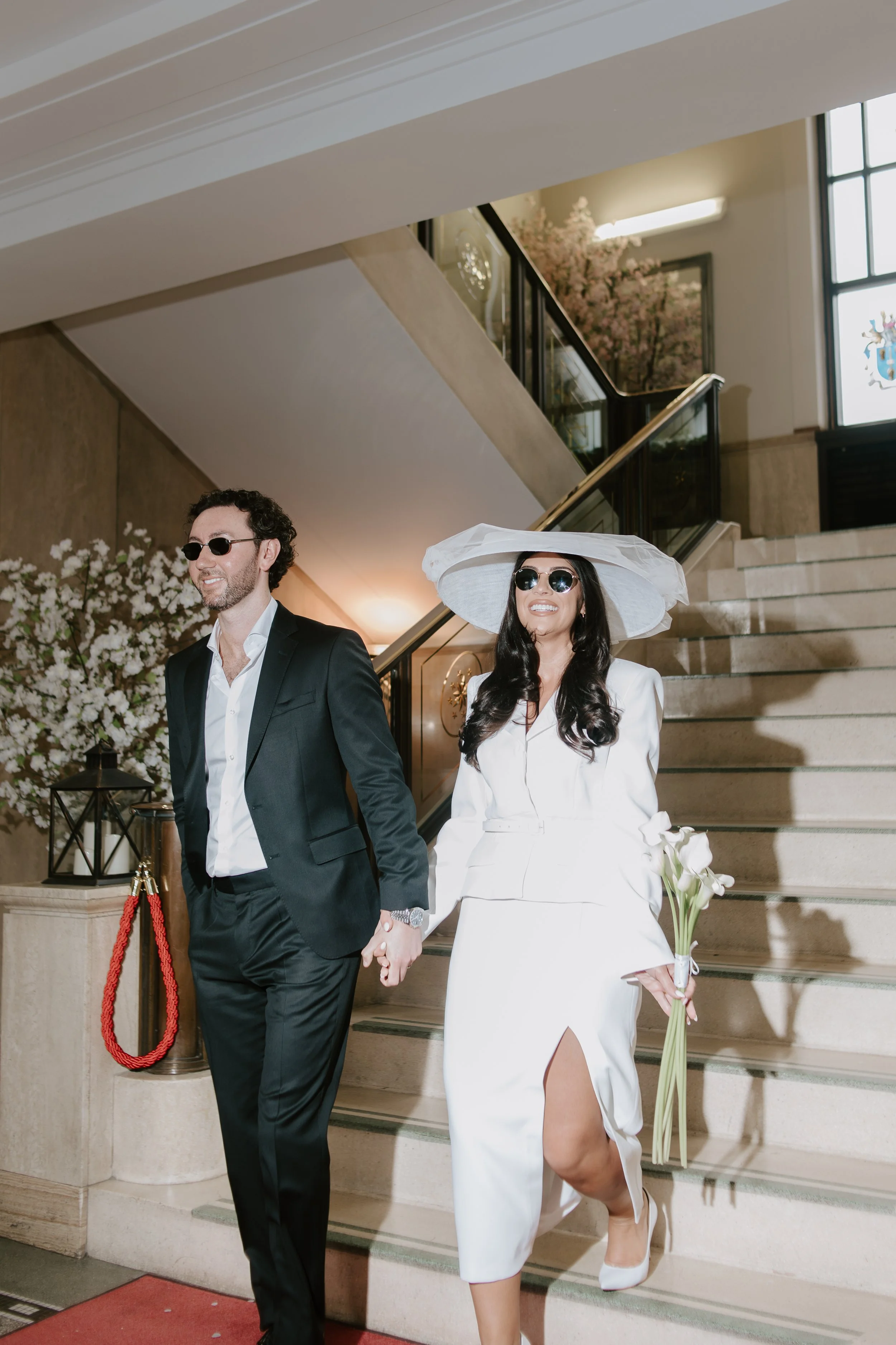 A man and woman in wedding attire walk down a staircase holding hands. The woman wears a white dress, large white hat, and sunglasses, holding a small bouquet, while the man wears a black suit and sunglasses. The setting appears to be indoors with de
