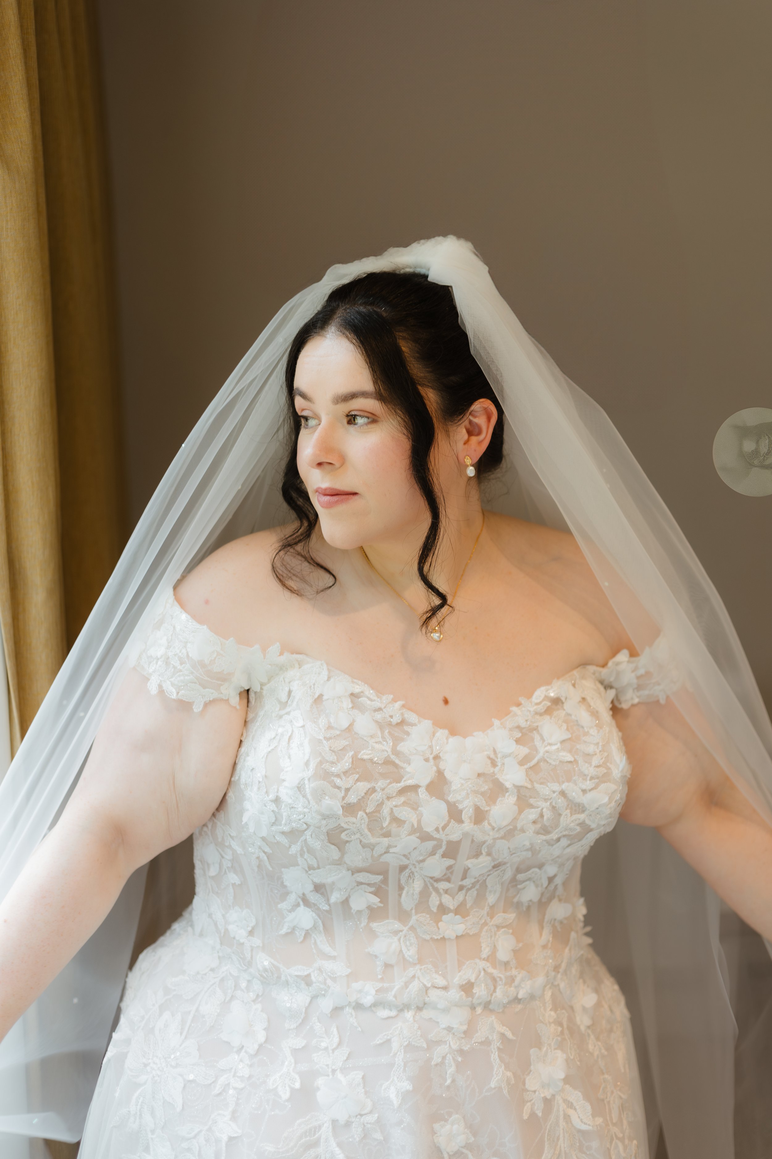 A bride in a white wedding dress with floral lace details, wearing a veil and pearl earrings, standing indoors by a window.