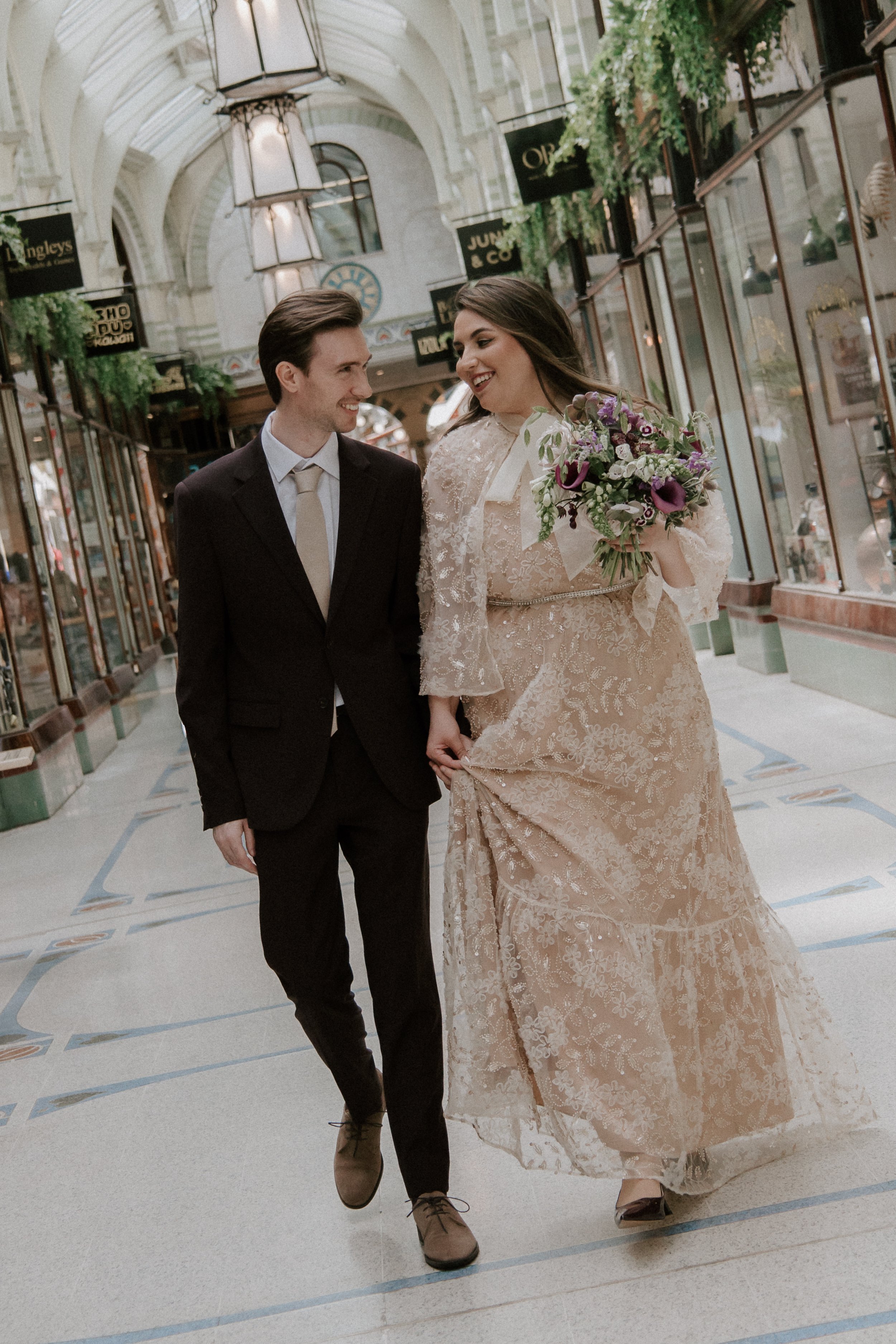 A couple dressed in wedding attire walking inside a shopping arcade, with the woman holding a bouquet of flowers and smiling at the man.