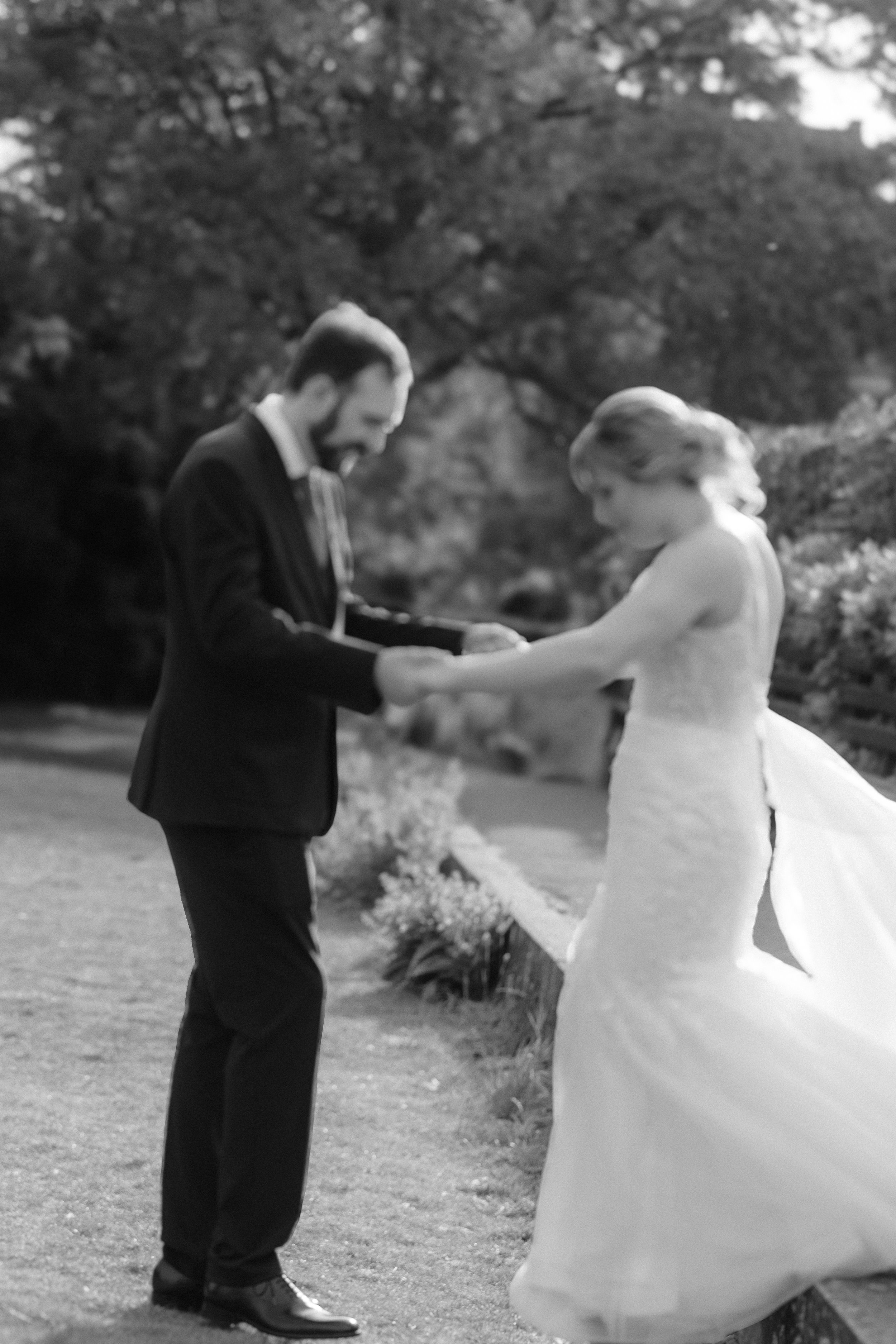 A black and white photo of a couple sharing a wedding moment outdoors, with the man holding the woman's hands as they look at each other.