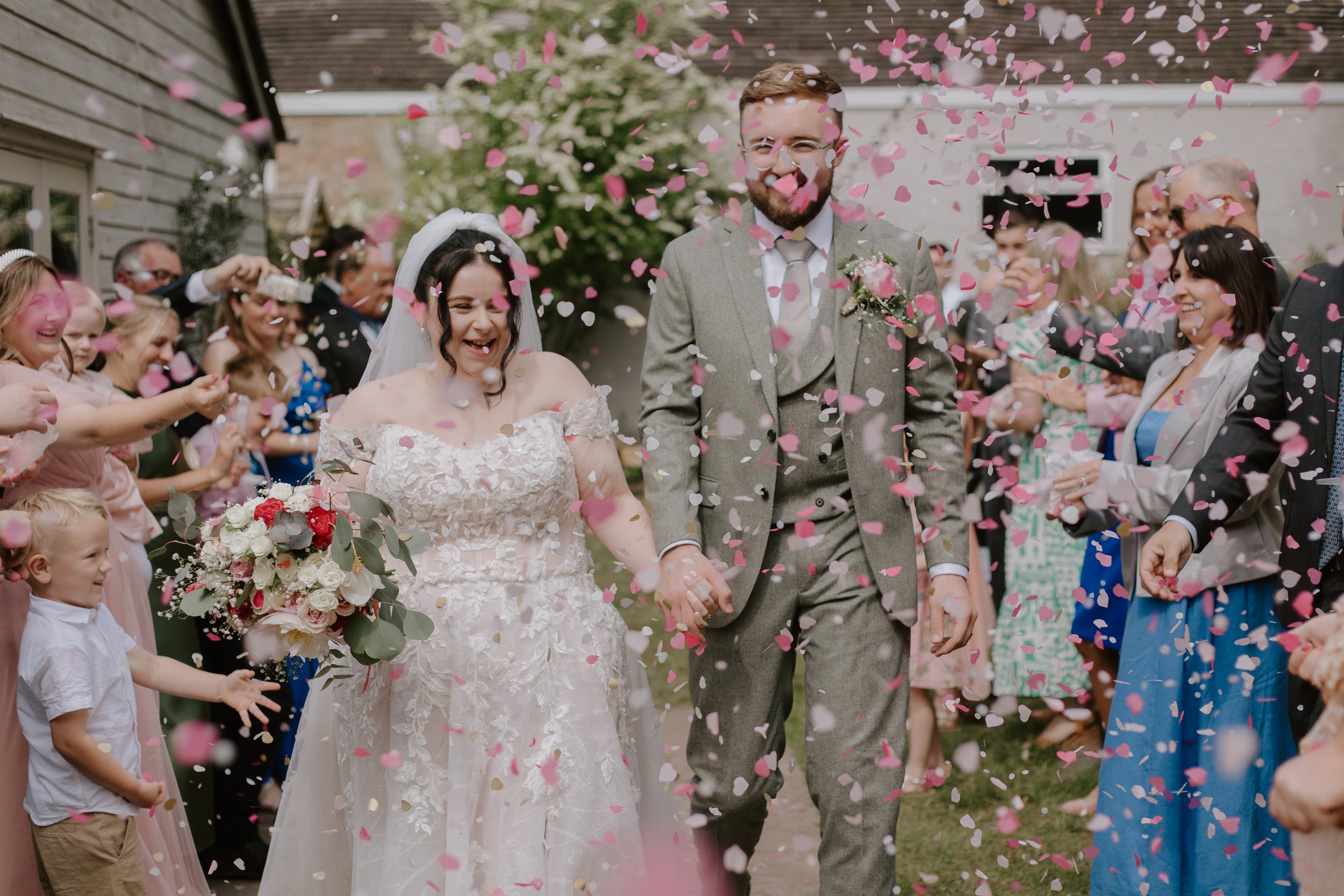 A newlywed couple holding hands and walking through a shower of pink and white confetti, surrounded by friends and family celebrating outdoors.