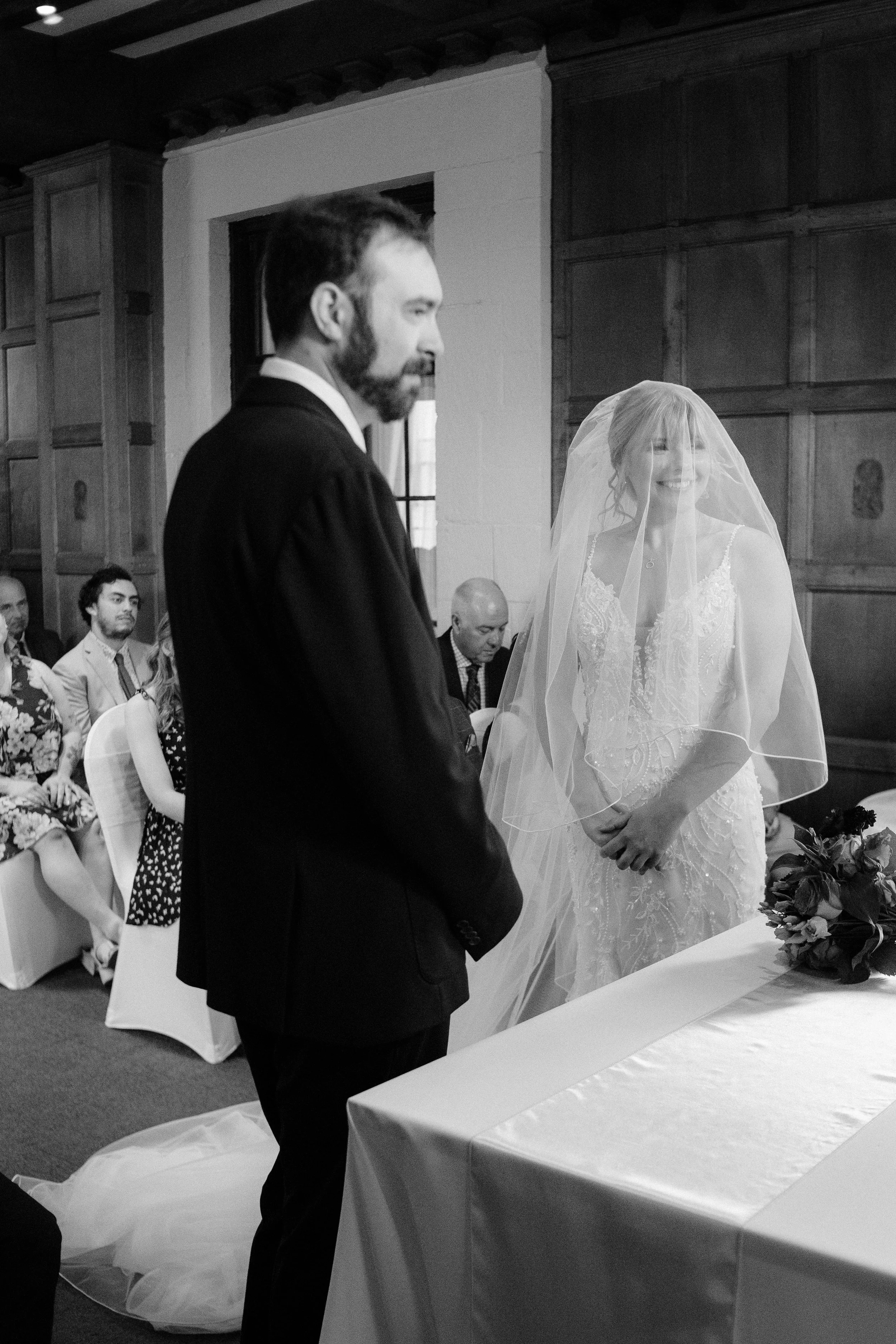 A black and white photo of a wedding ceremony with a bride and groom standing before an officiant. The bride is smiling, wearing a lace wedding gown and veil, while the groom has a beard and is dressed in a dark suit. They are indoors, with guests se