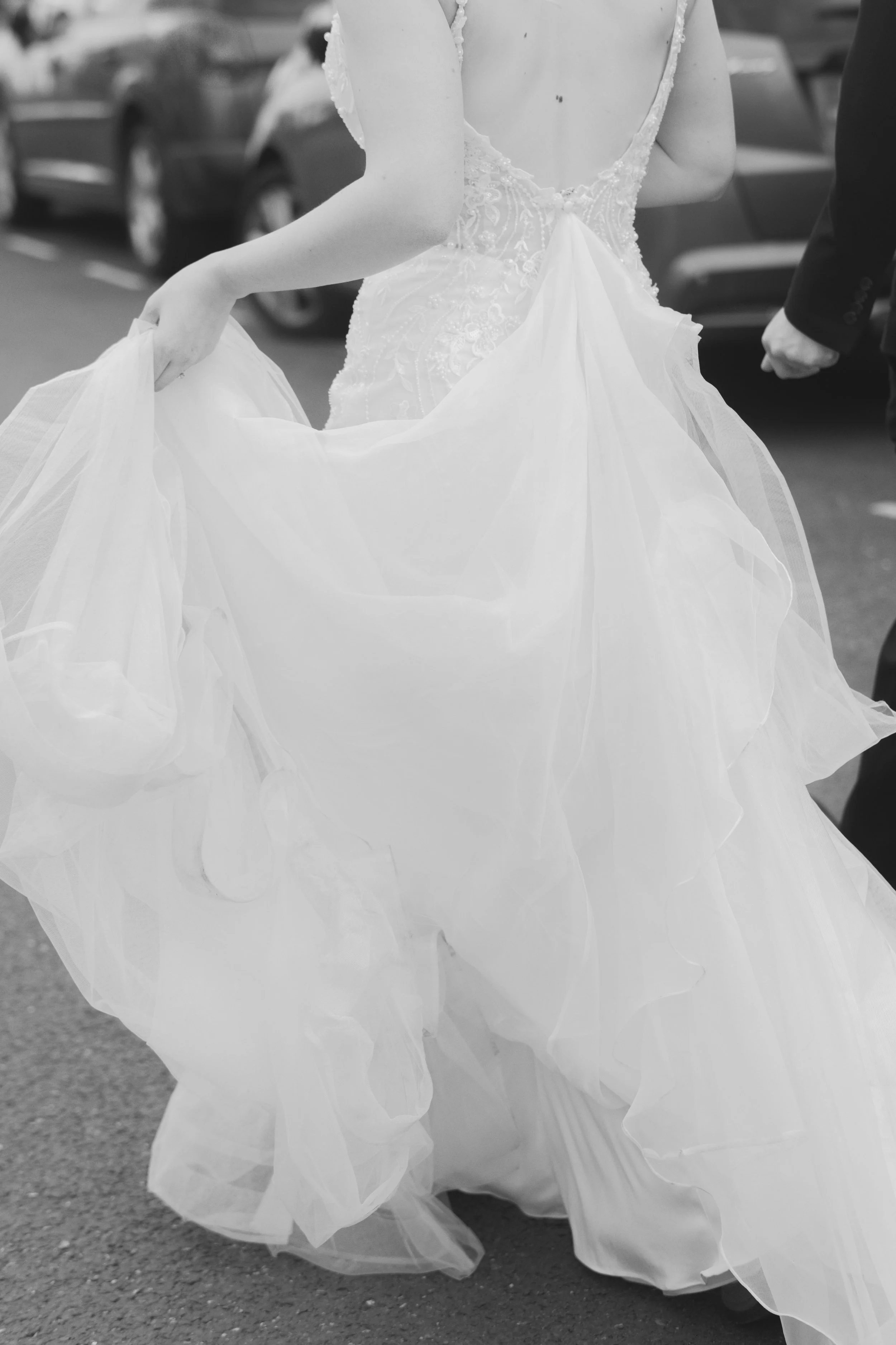 Black and white photo of a bride holding up her wedding dress, standing on a parking lot with cars in the background.