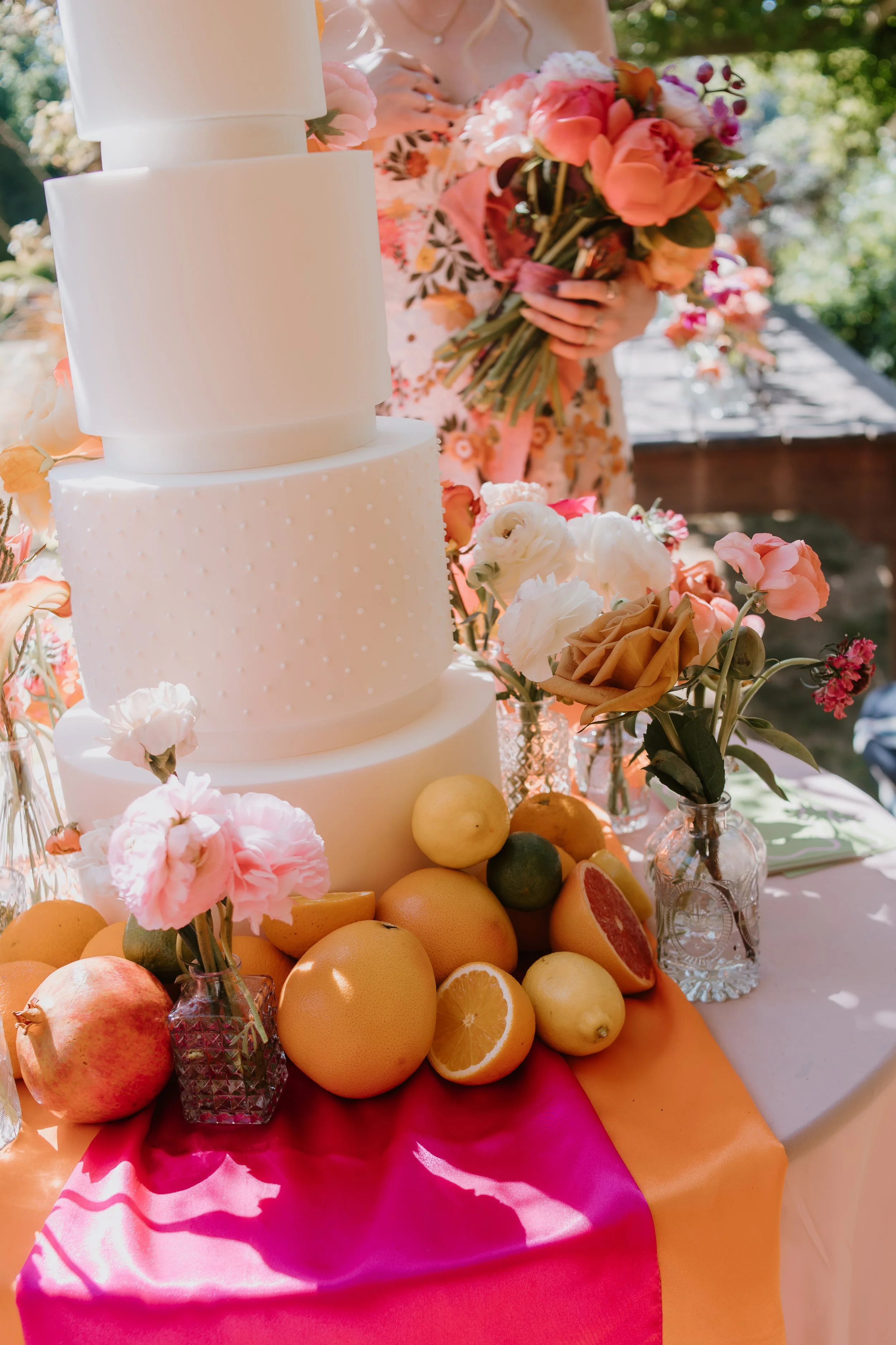 A four-tier white wedding cake with textured icing, surrounded by assorted fresh fruits and flowers on a colorful tablecloth, with a bride holding a bouquet in the background.