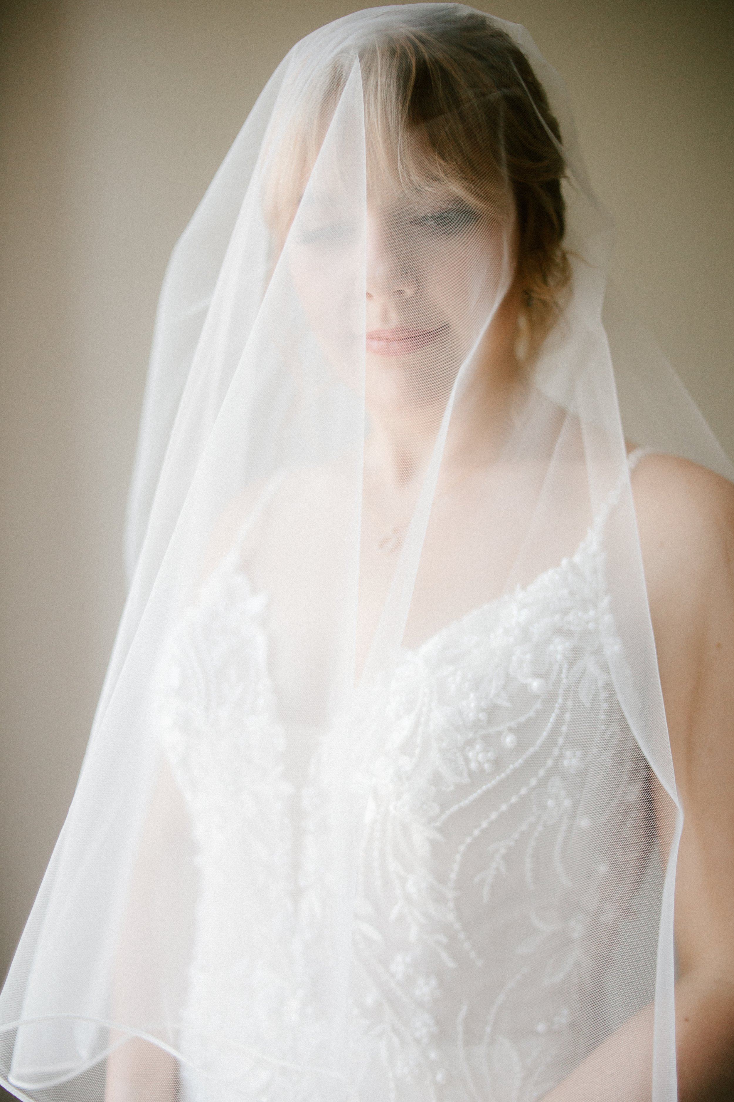 A bride with a short hairstyle wearing a wedding dress and veil, standing in a softly lit room.