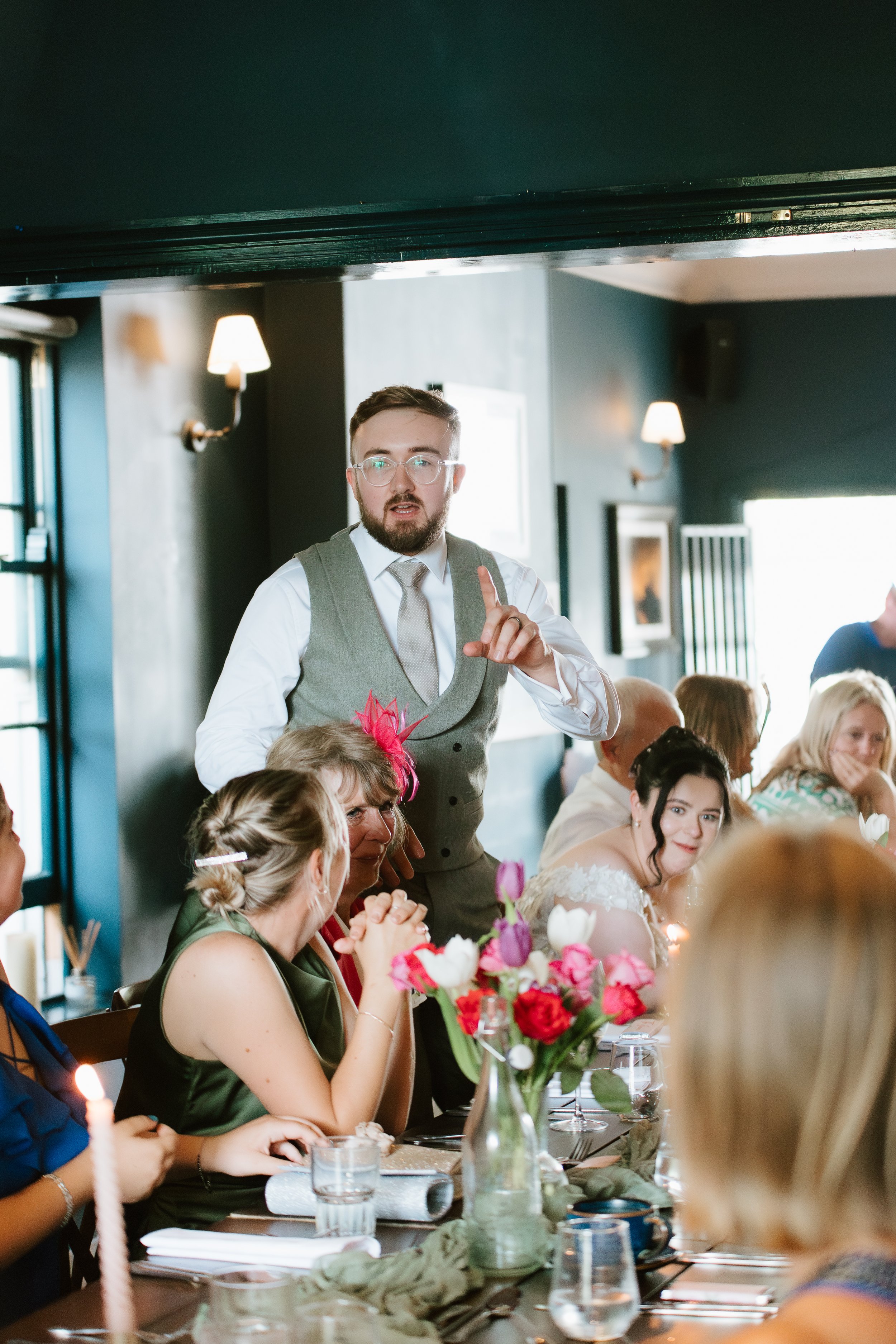A man in a gray vest and white shirt appears to be giving a speech at a wedding reception, with women seated at a decorated table in front of him.