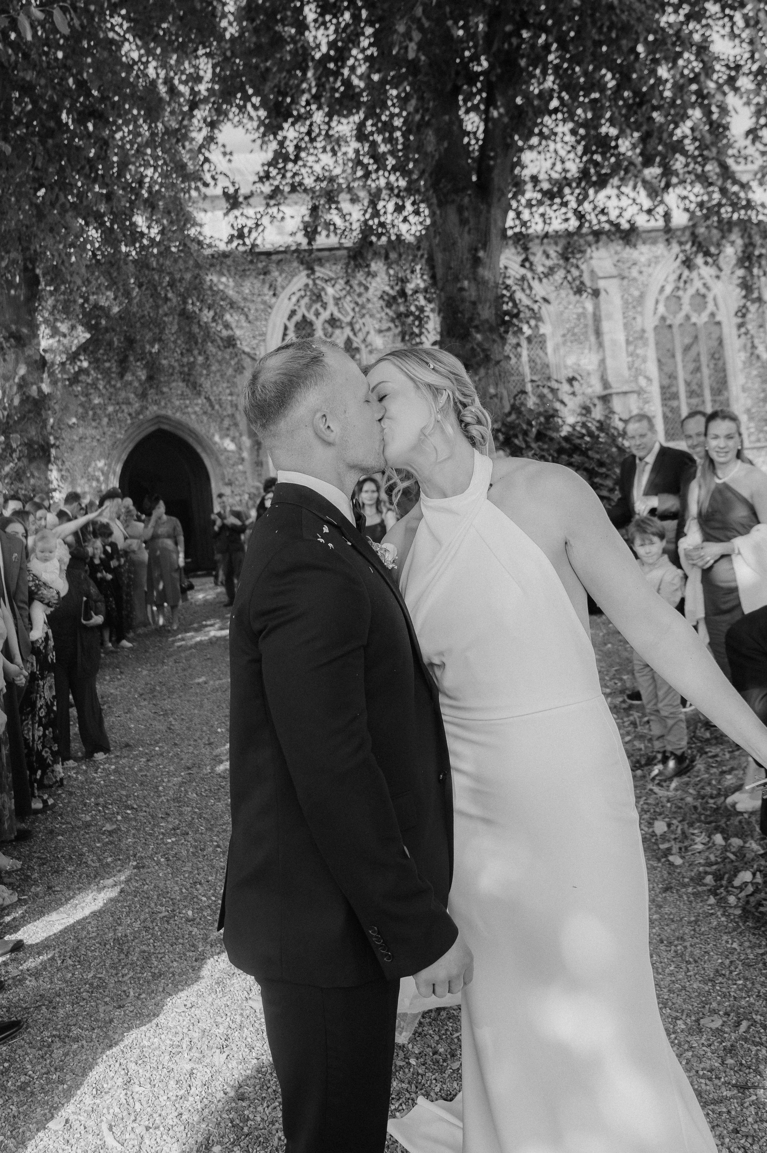 Timeless and documentary wedding photographer Suffolk and Norfolk – black and white photo of newlywed couple kissing outside church surrounded by guests