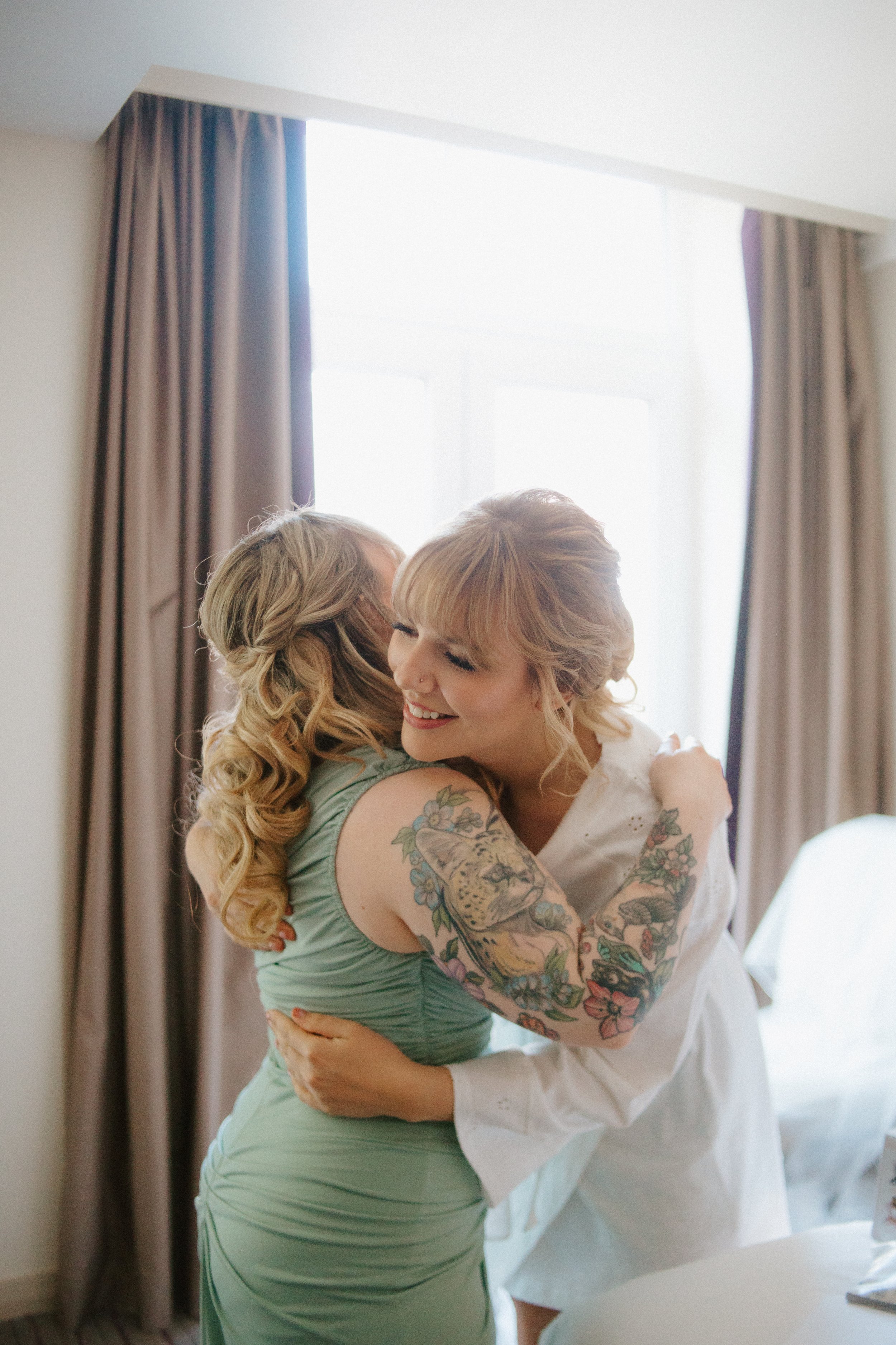 Two women hugging and smiling in a bright room, one wearing a light green dress and the other in a white dress with tattoos on her arms.