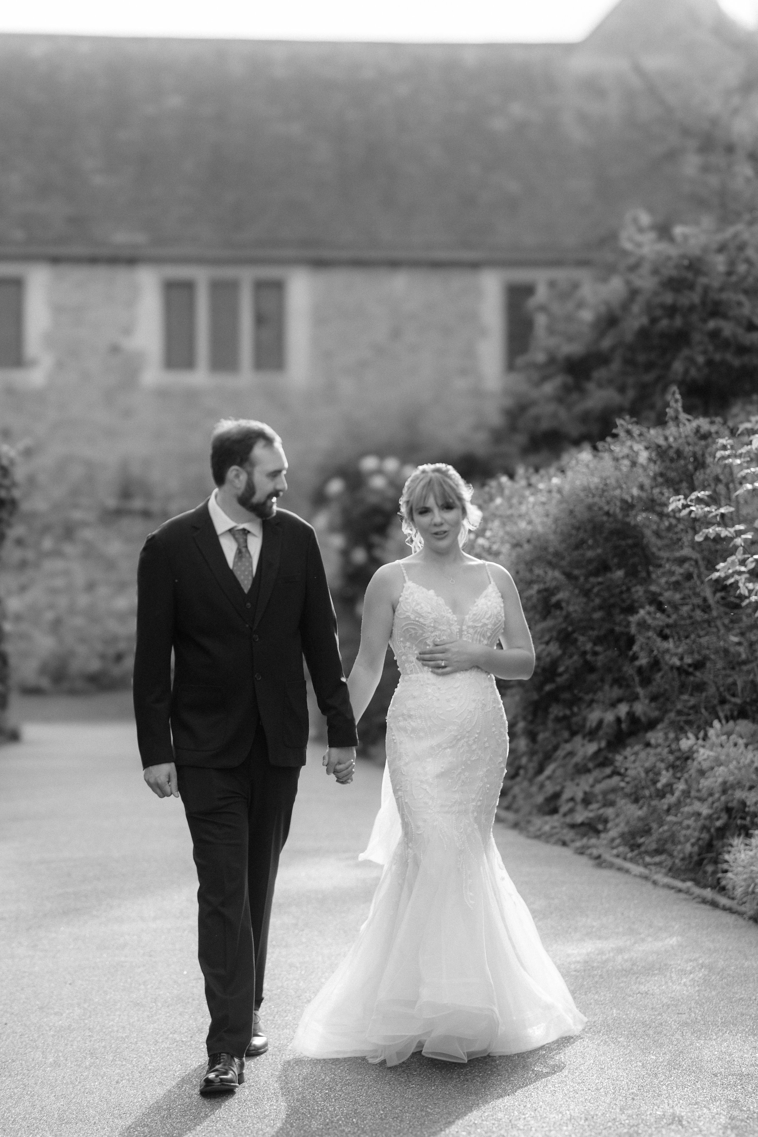 A black and white photograph of a bride and groom walking hand-in-hand outdoors. The bride is in a lace wedding gown, and the groom is in a dark suit and tie. They are surrounded by greenery and a large stone building in the background.