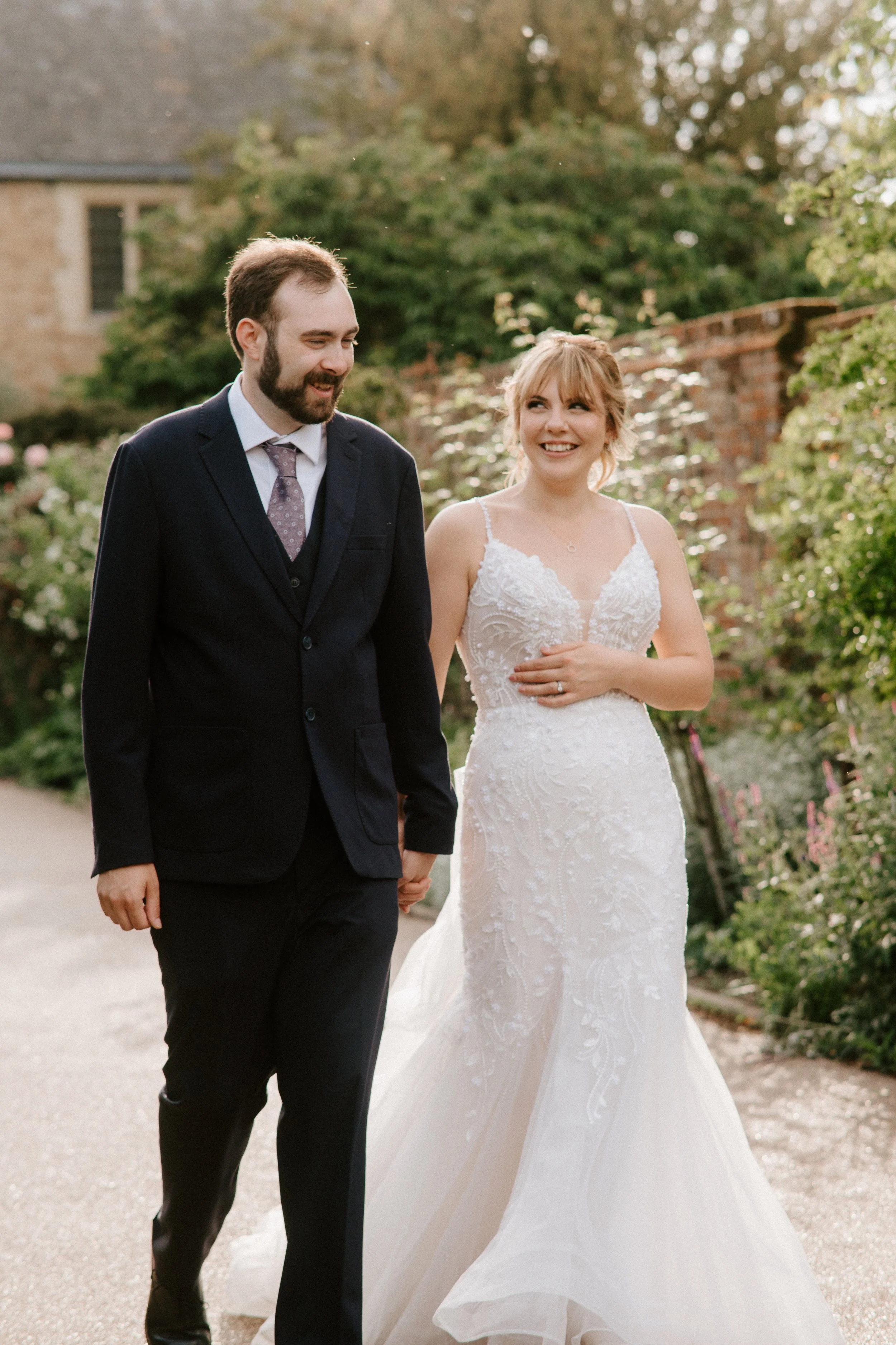 A newlywed couple walking outdoors, holding hands. The bride is wearing a white lace wedding gown, and the groom is in a dark suit with a patterned tie. They are smiling and looking at each other amid greenery and a brick wall in the background.