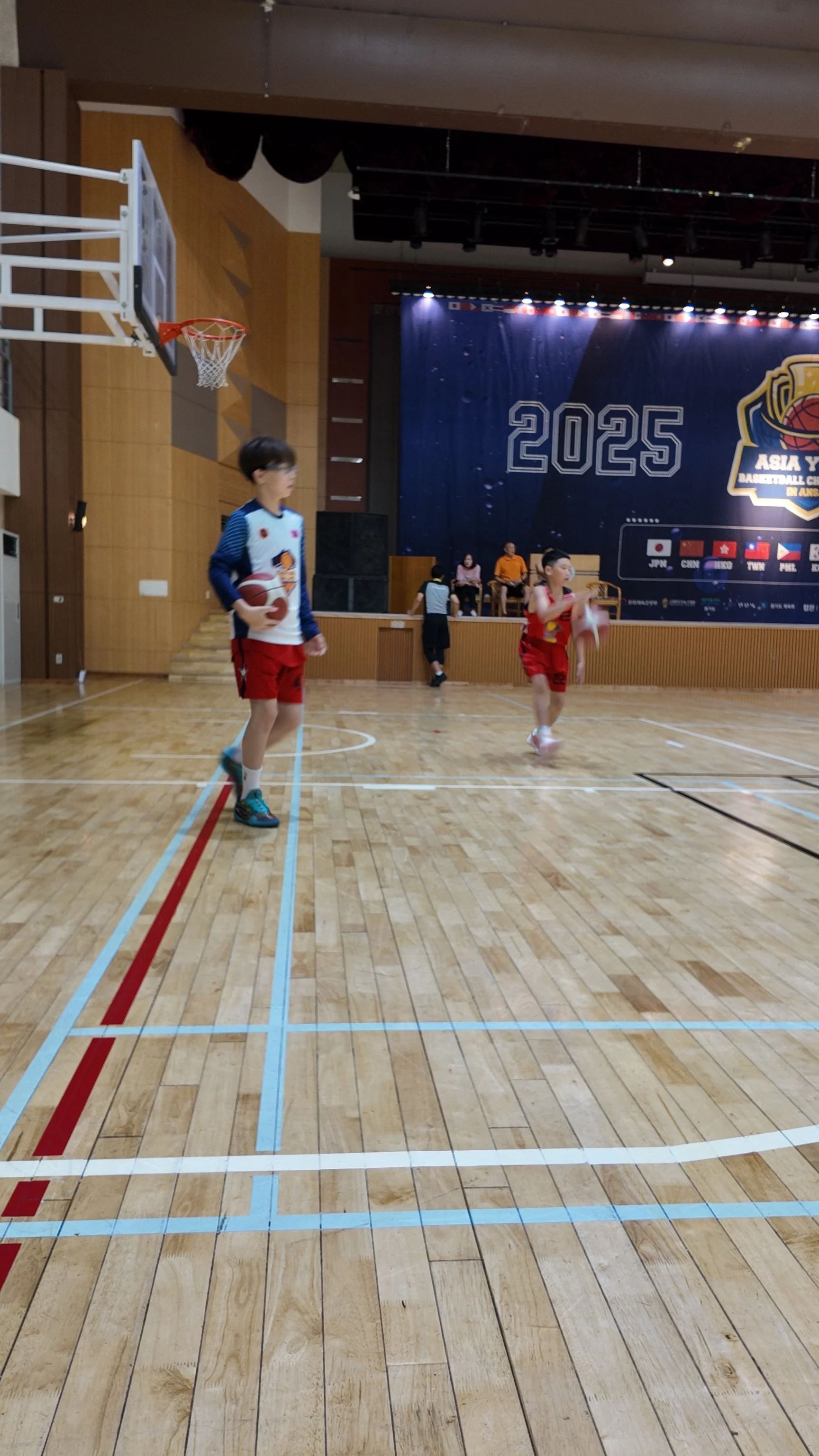 Two children practicing basketball on an indoor court, one holding a basketball and the other preparing to shoot, with a stage and a large banner in the background.